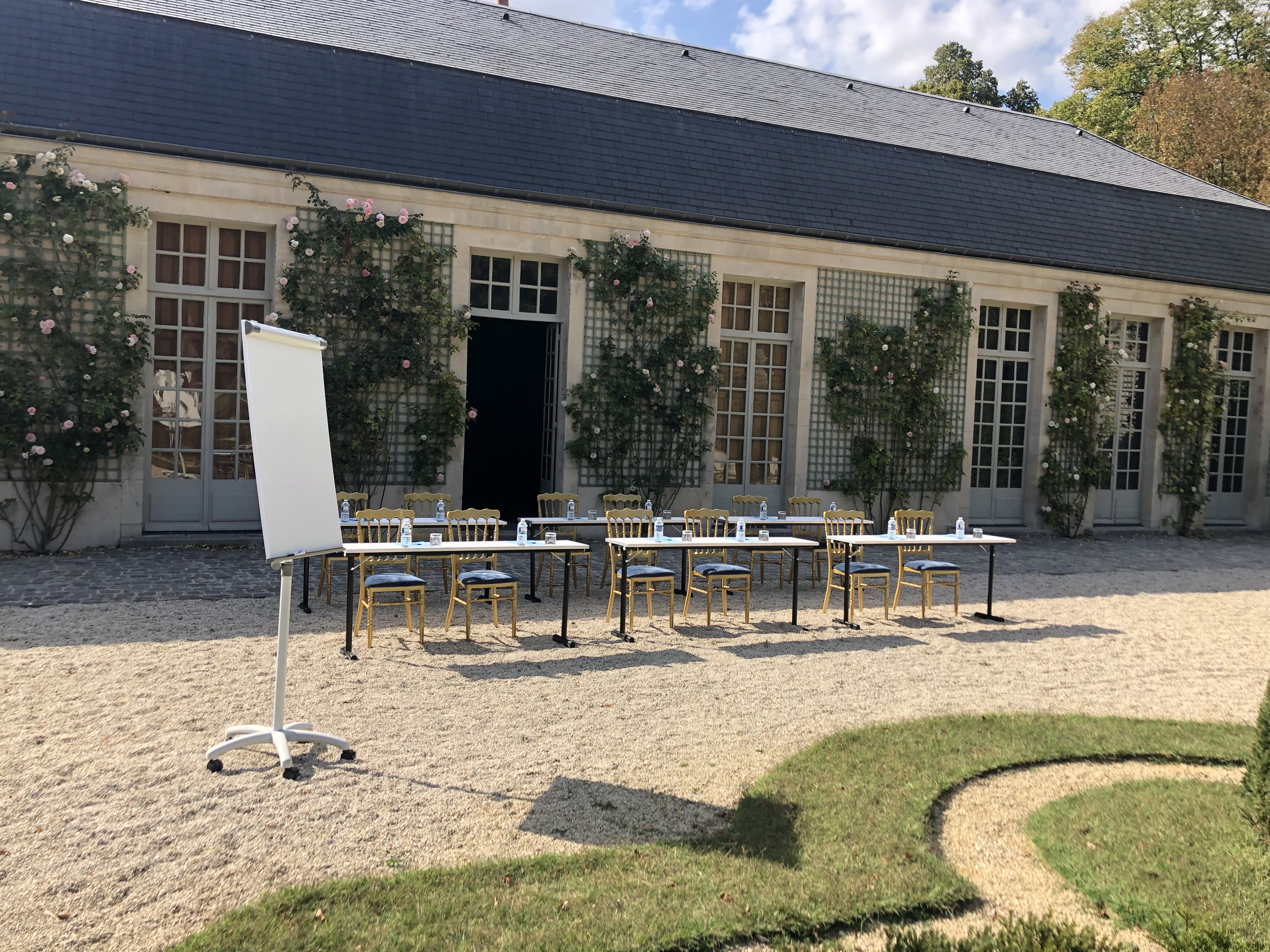 Outdoor seminar setup with tables, chairs, bottled water, and a flip chart in front of a building with tall windows and climbing roses.