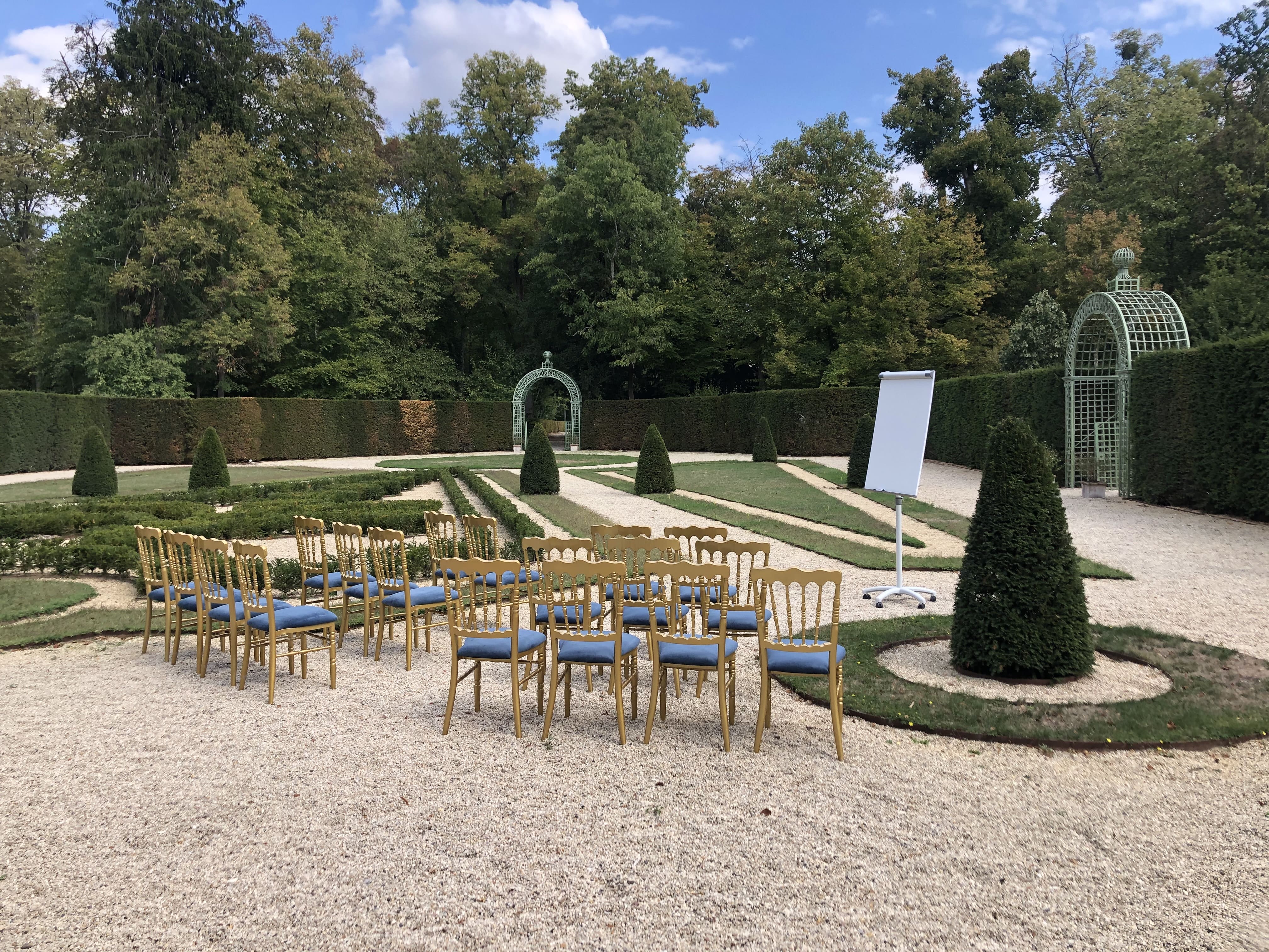 Outdoor garden seminar setup with gold chairs with blue cushions arranged facing a blank flip chart, surrounded by manicured hedges and green trees under a partly cloudy sky.