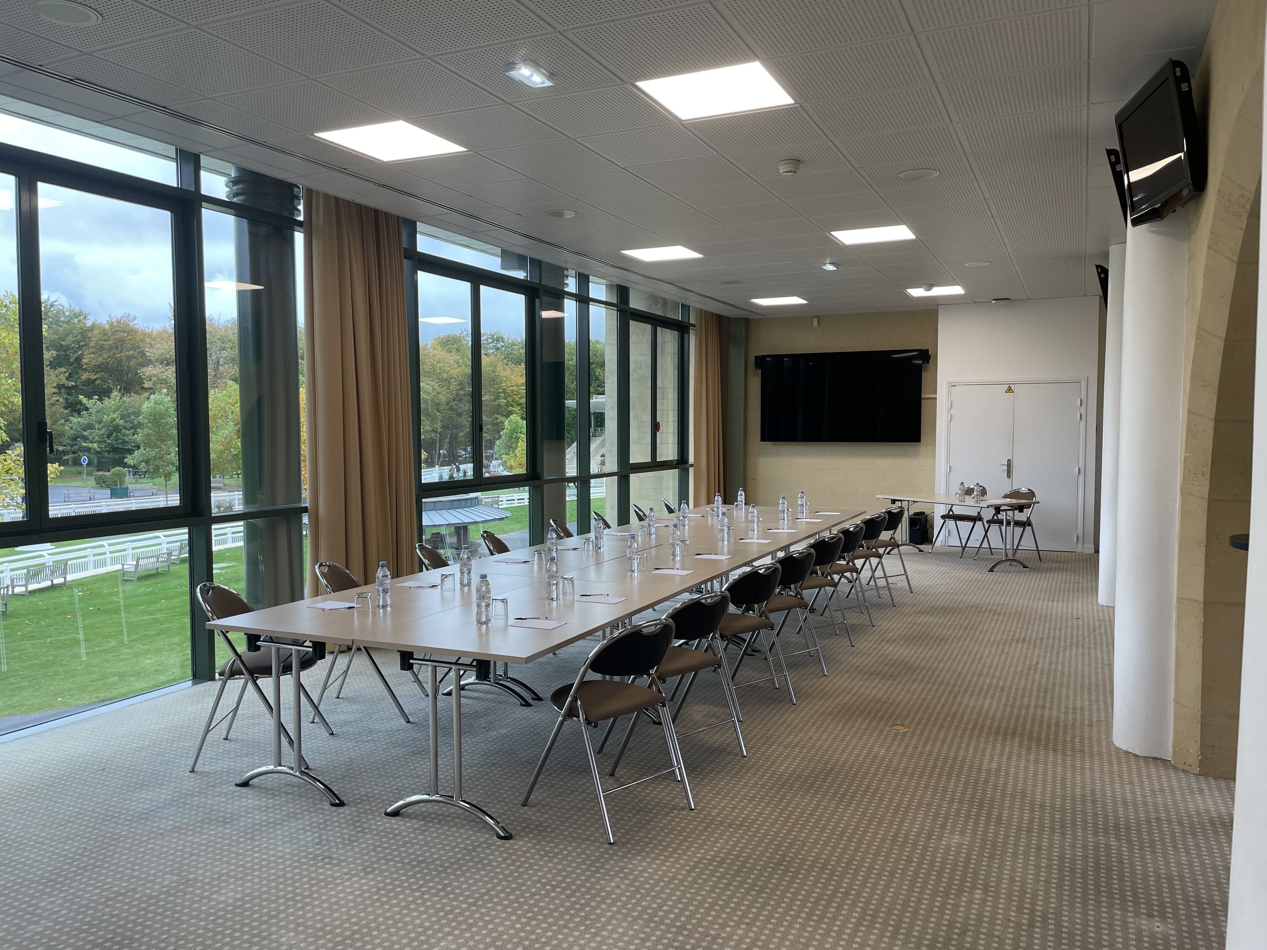 Long conference table with chairs, water bottles, and notepads in a bright meeting room with large windows overlooking green trees.