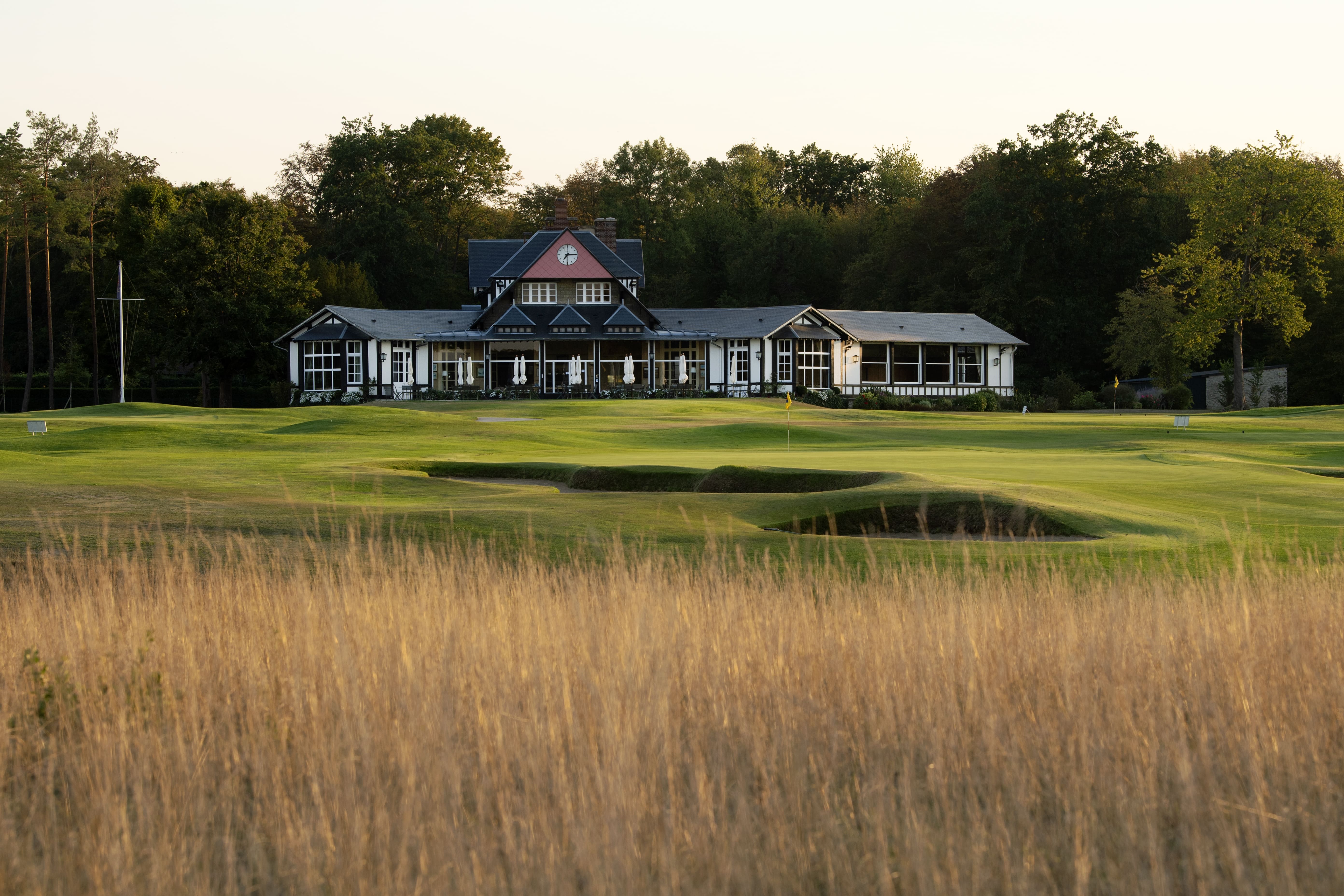 Golf course green with sand bunkers in front of a large clubhouse surrounded by trees.