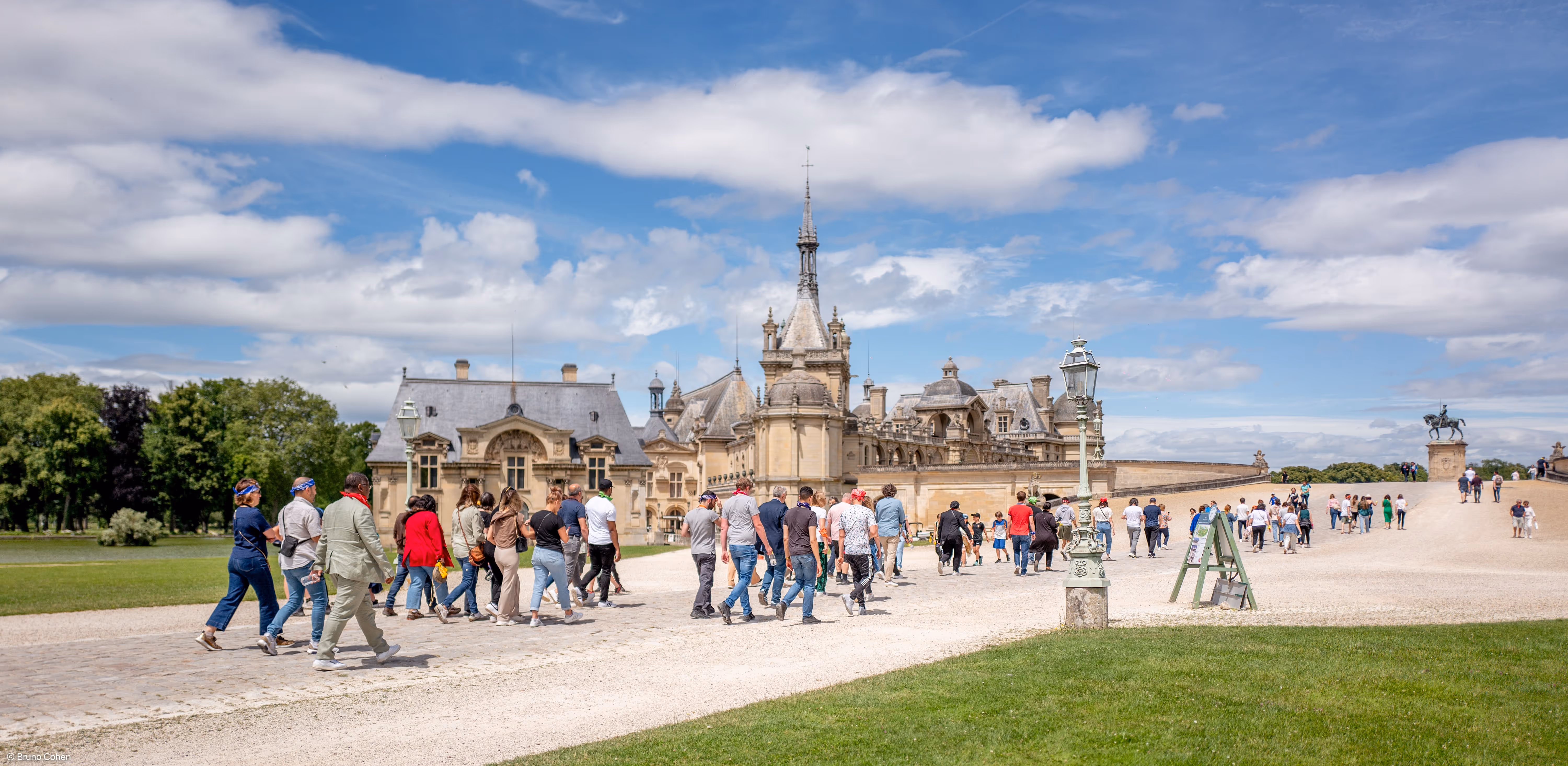Group of people walking on a pathway near a historic chateau under a partly cloudy sky.