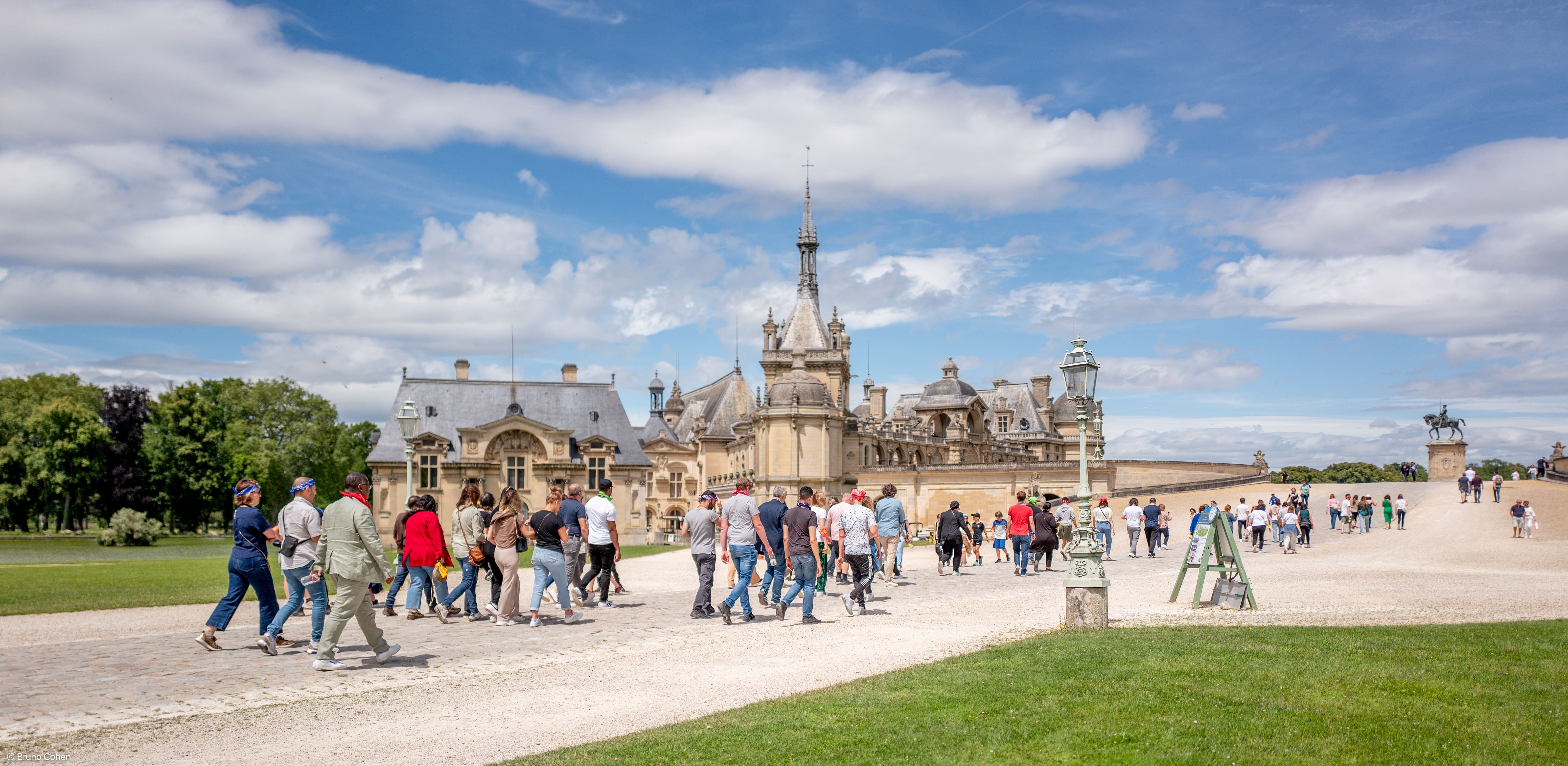 Group of people walking on a pathway near a historic chateau under a partly cloudy sky.