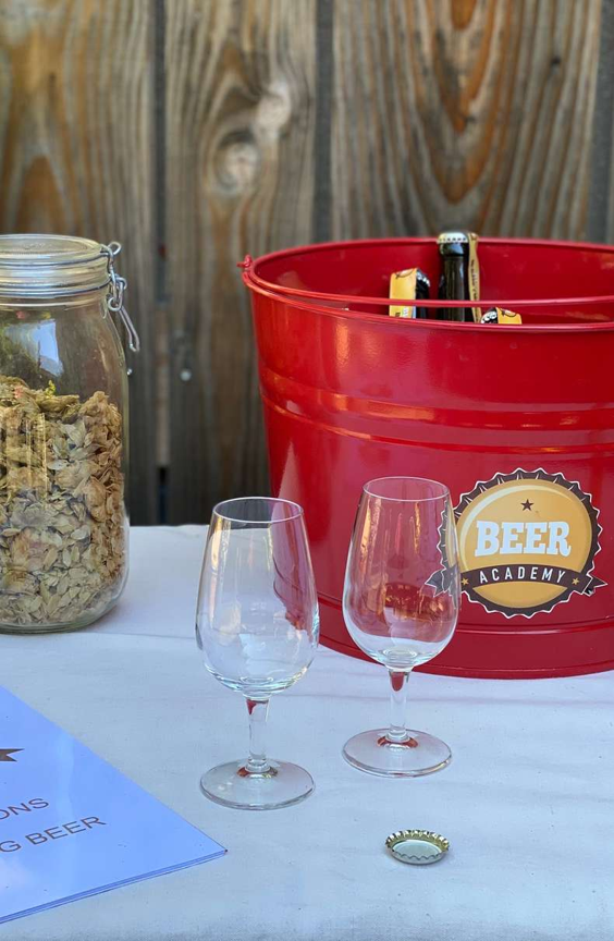 Red bucket with bottled beers, two empty beer glasses, a jar filled with dried hops, and a blue booklet on a white table.