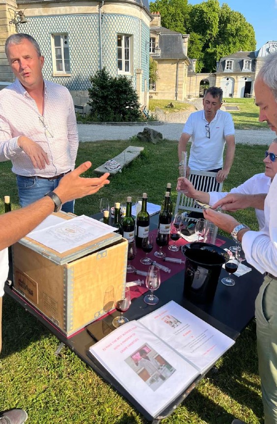 Group of men outdoors around a table with wine bottles, glasses, and documents, appearing to conduct a wine tasting near a historic building.
