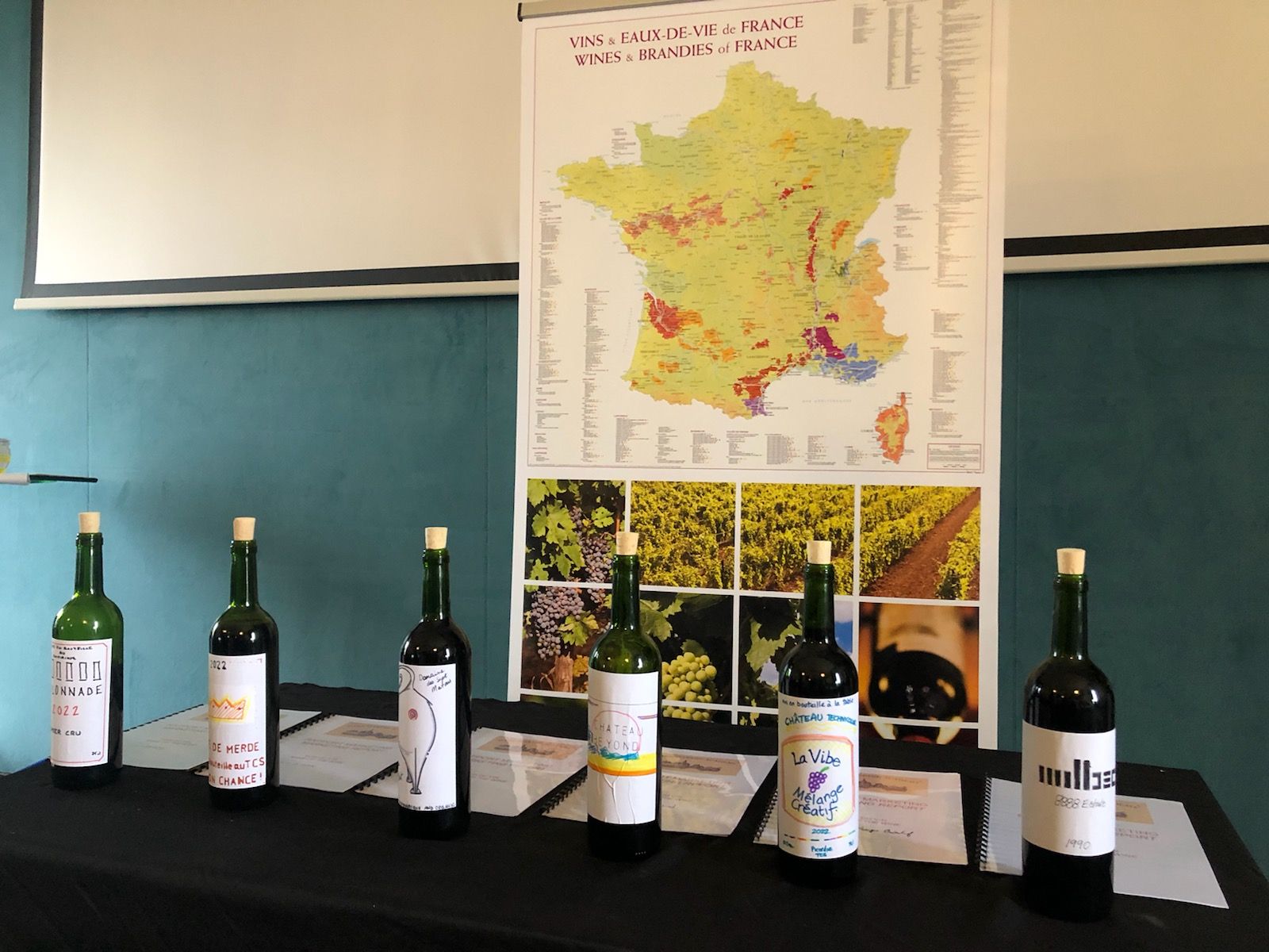 Six wine bottles with handwritten labels on a black table in front of a colorful map of French wine regions and photos of grapevines.