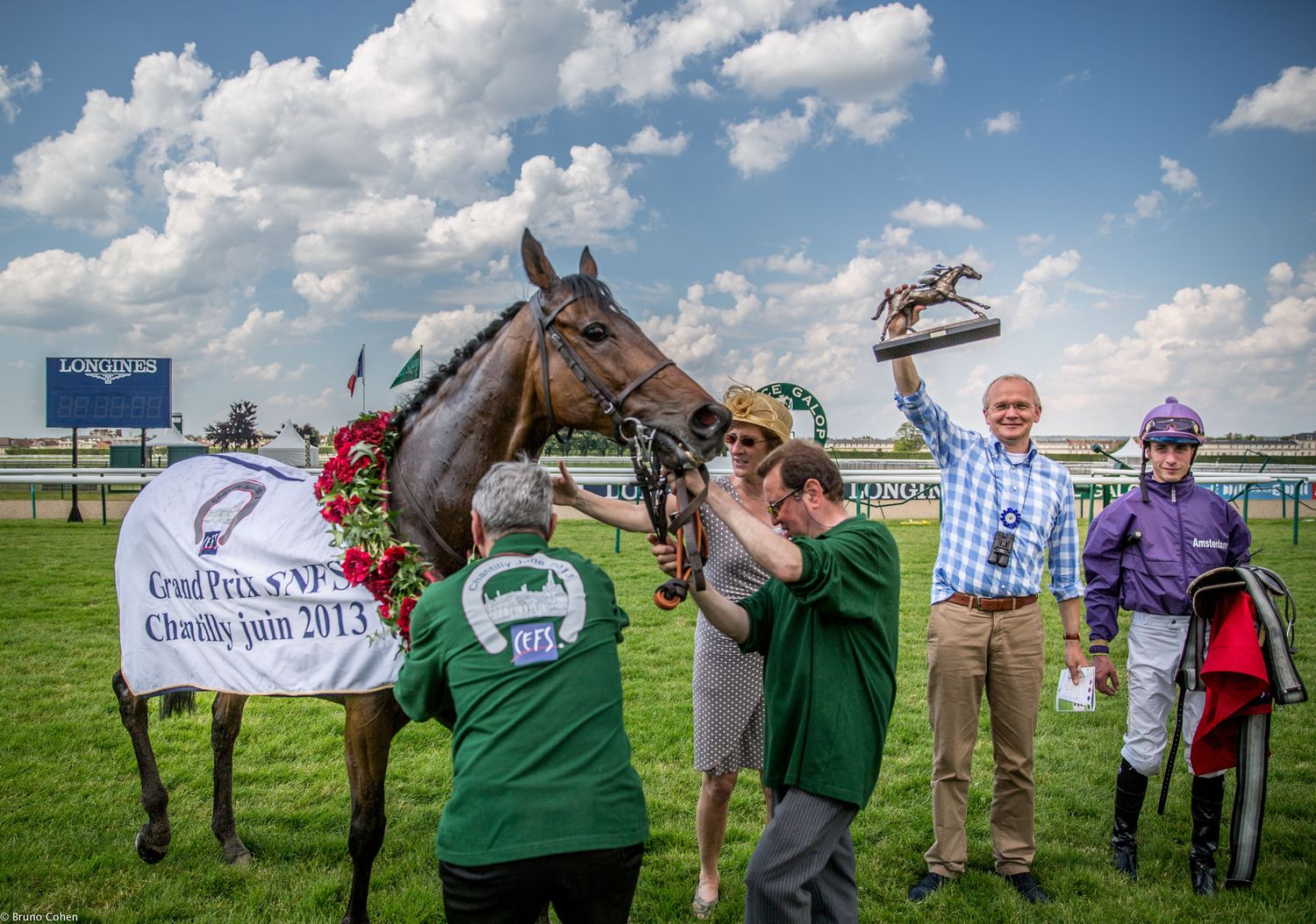 Racehorse adorned with a floral garland and a winner's blanket, surrounded by four people celebrating on a racetrack.