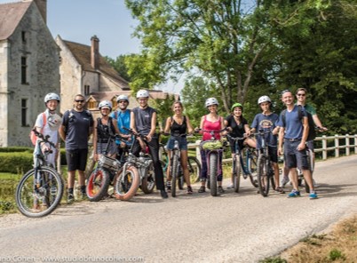 Group of people standing with electric bicycles on a sunny roadside near trees and old stone buildings.