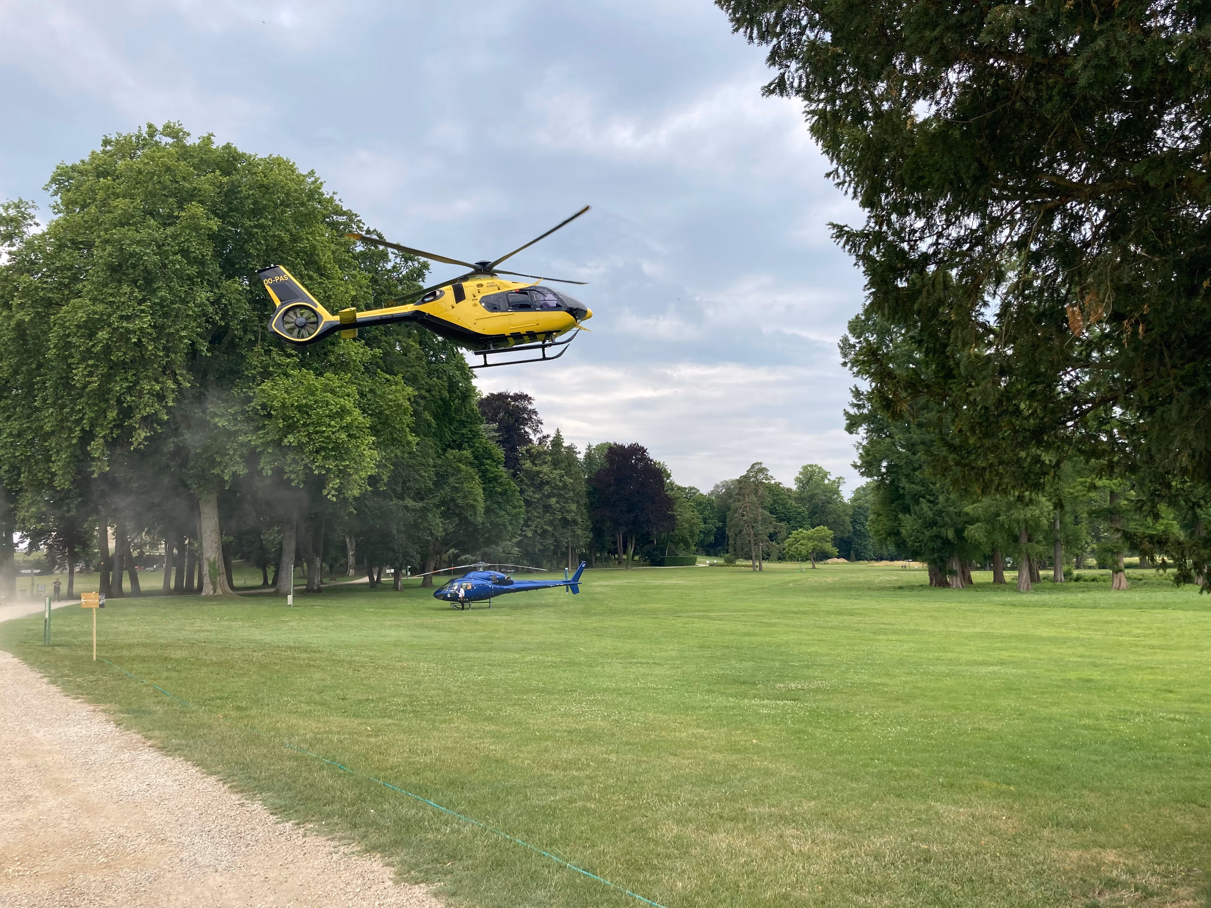 Yellow helicopter hovering above grass near a blue helicopter parked on a green lawn surrounded by trees.