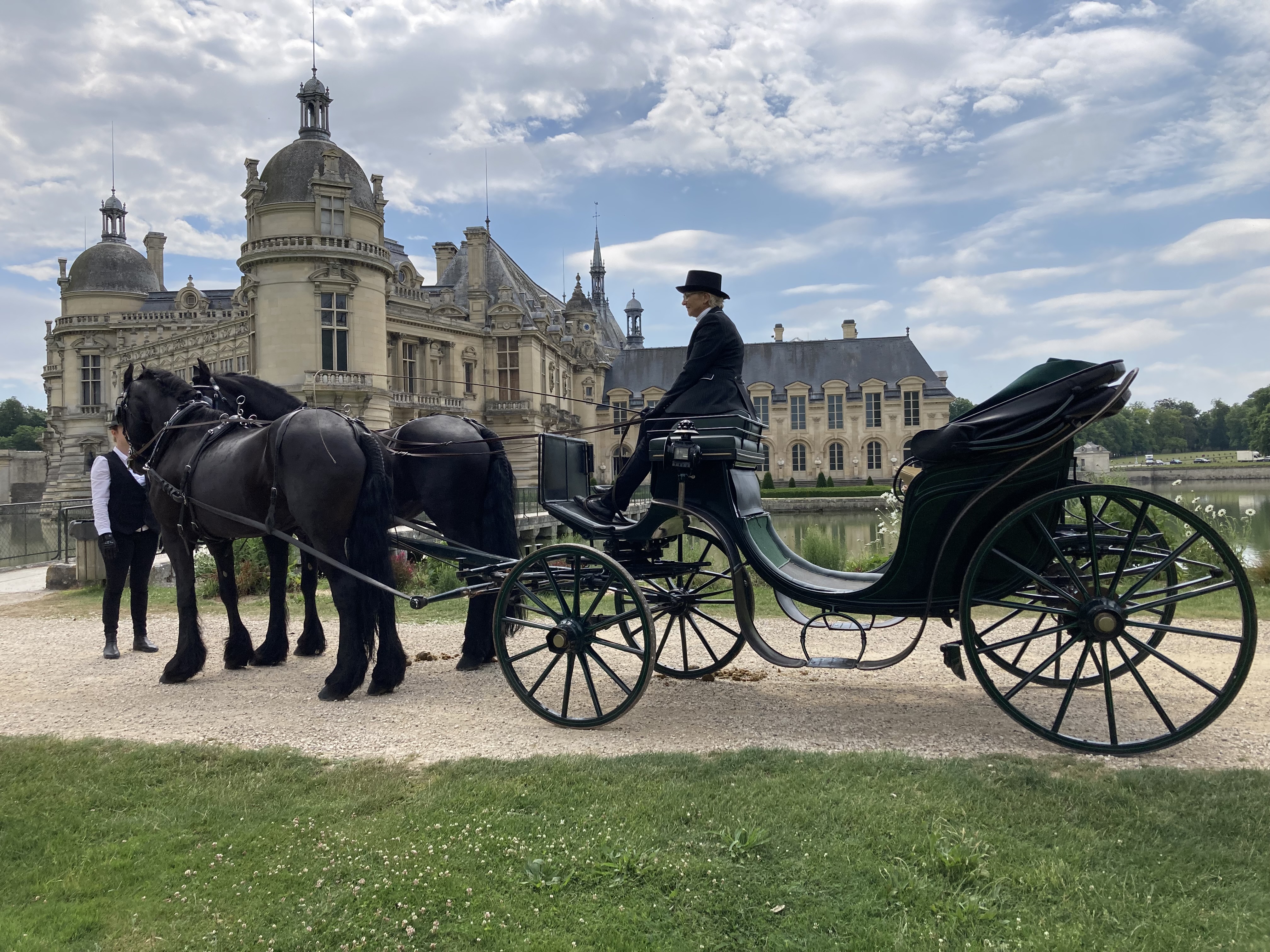 Two black horses hitched to a vintage carriage with a driver in formal attire in front of a historic chateau under a partly cloudy sky.