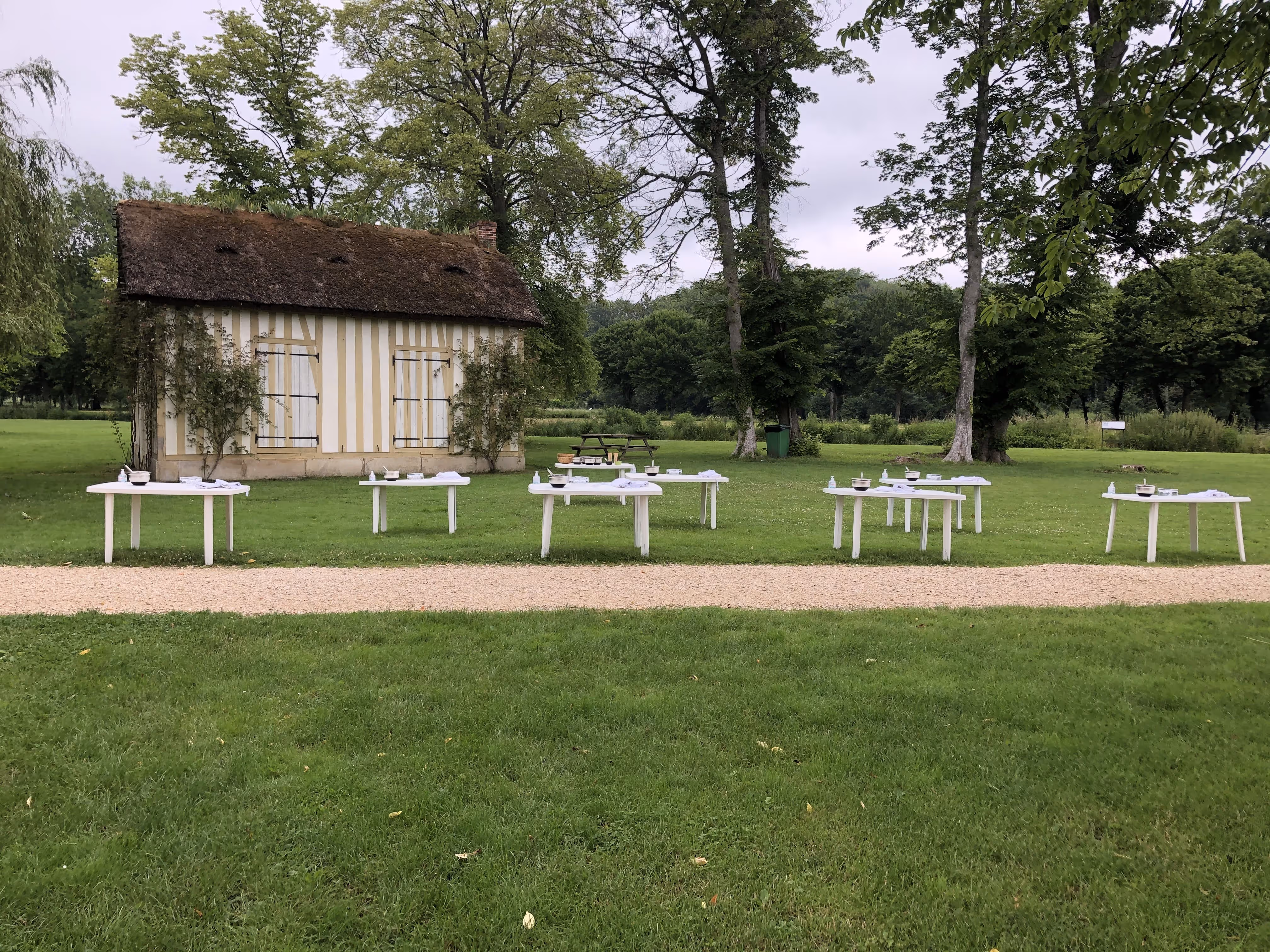 Small thatched-roof cottage with six white tables arranged outdoors on grass, each table set with bowls and utensils.
