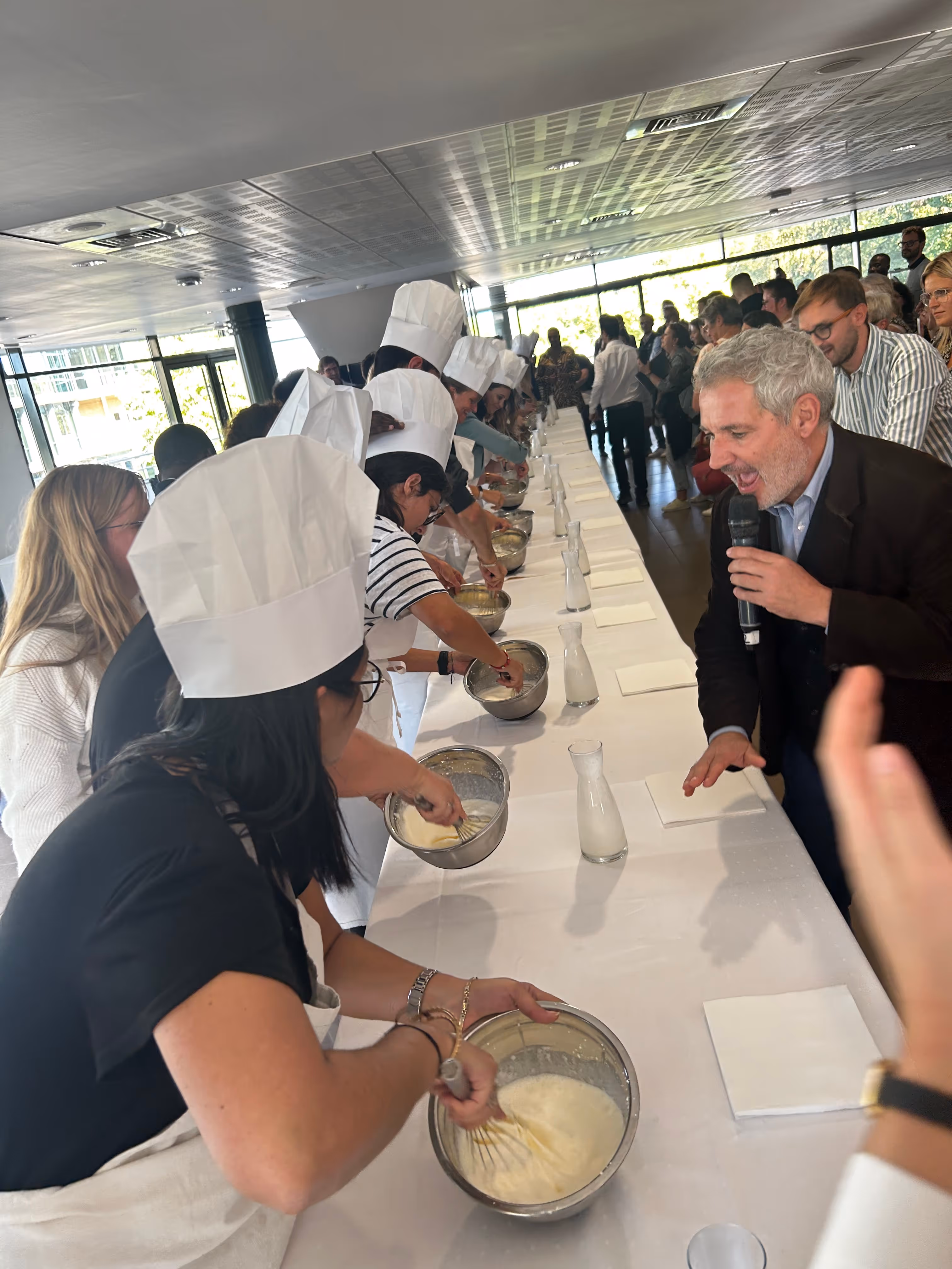 Several people wearing chef hats and aprons stand along a long table whisking mixtures in metal bowls while a man with gray hair speaks into a microphone.