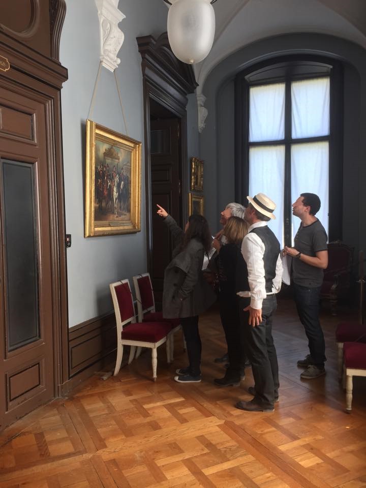 A group of five people standing in a museum room looking at and discussing a framed historical painting on the wall.