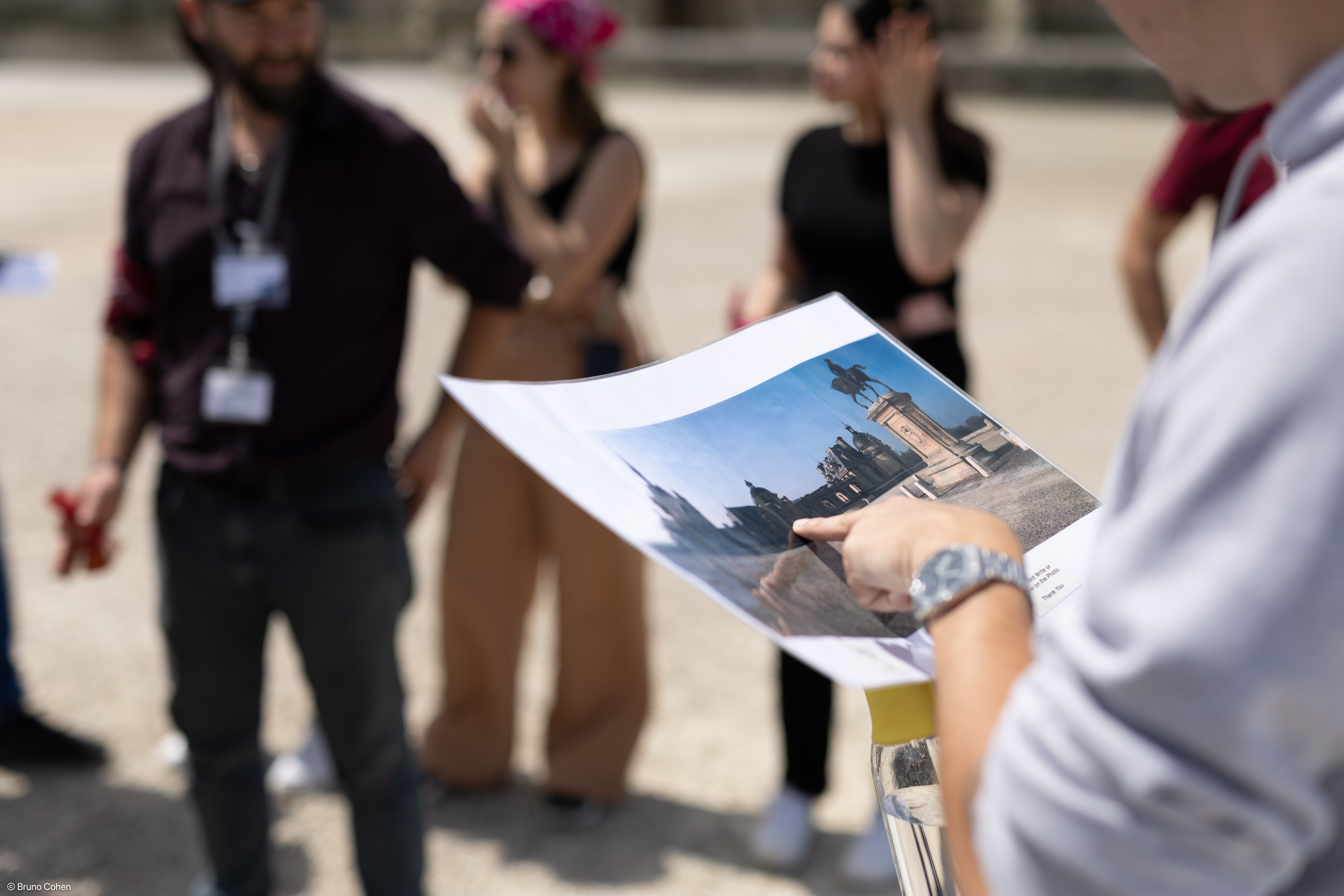 Person holding and pointing at a photo of a statue and buildings, with a group of people blurred in the background outdoors.