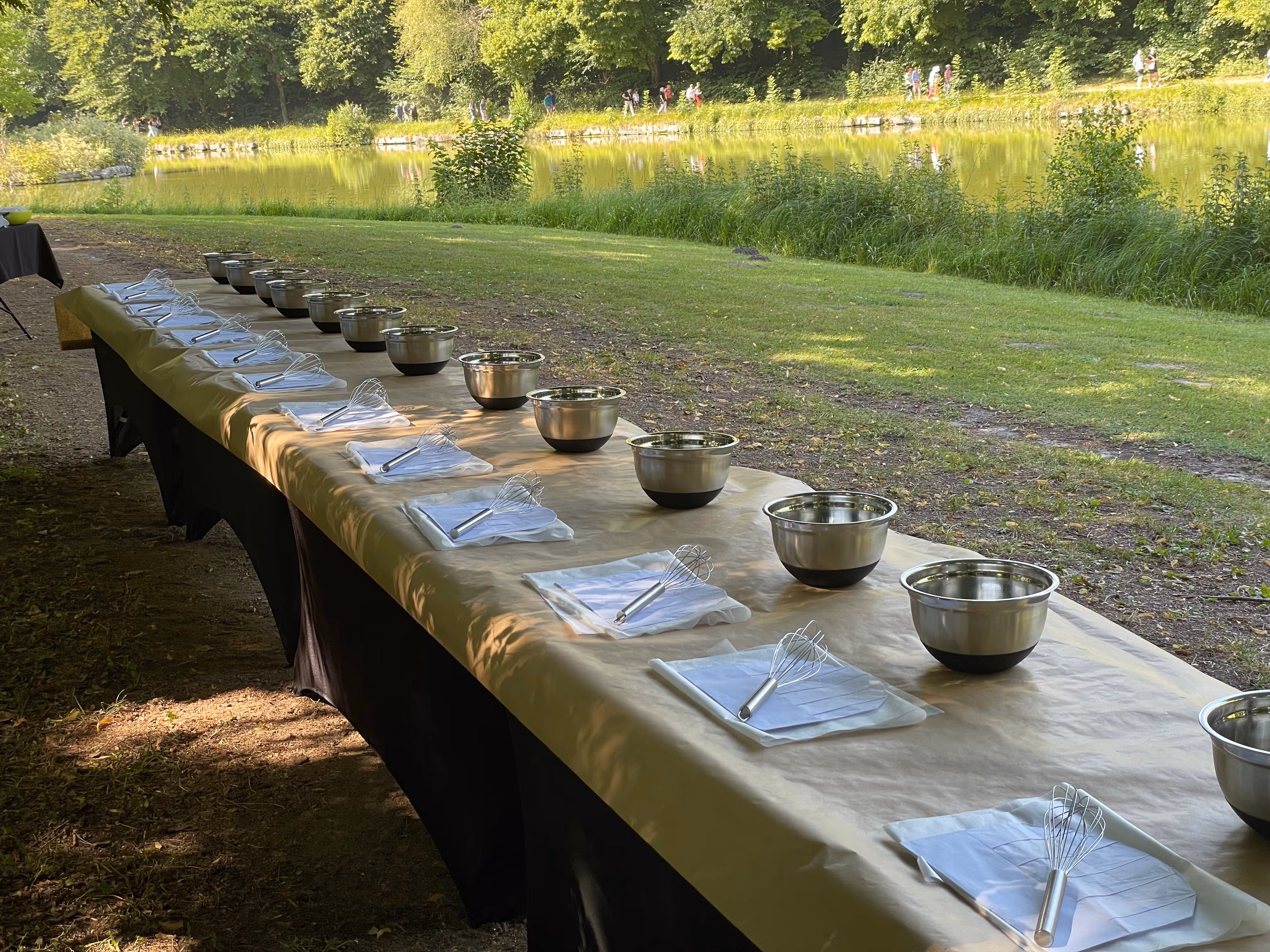 Long outdoor table lined with metal mixing bowls and whisks on folded napkins, set up near a pond and greenery.
