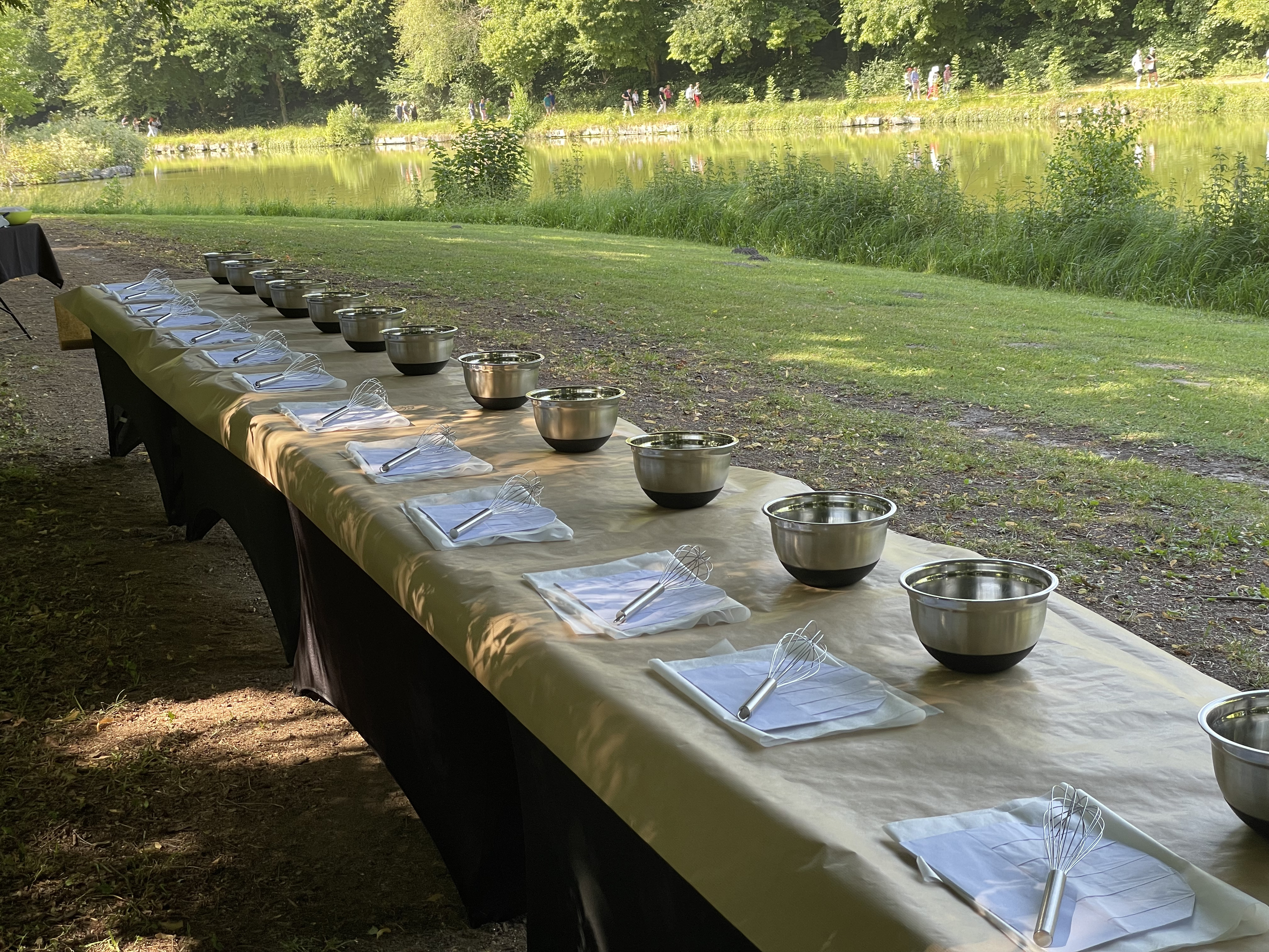 Long outdoor table lined with metal mixing bowls and whisks on folded napkins, set up near a pond and greenery.