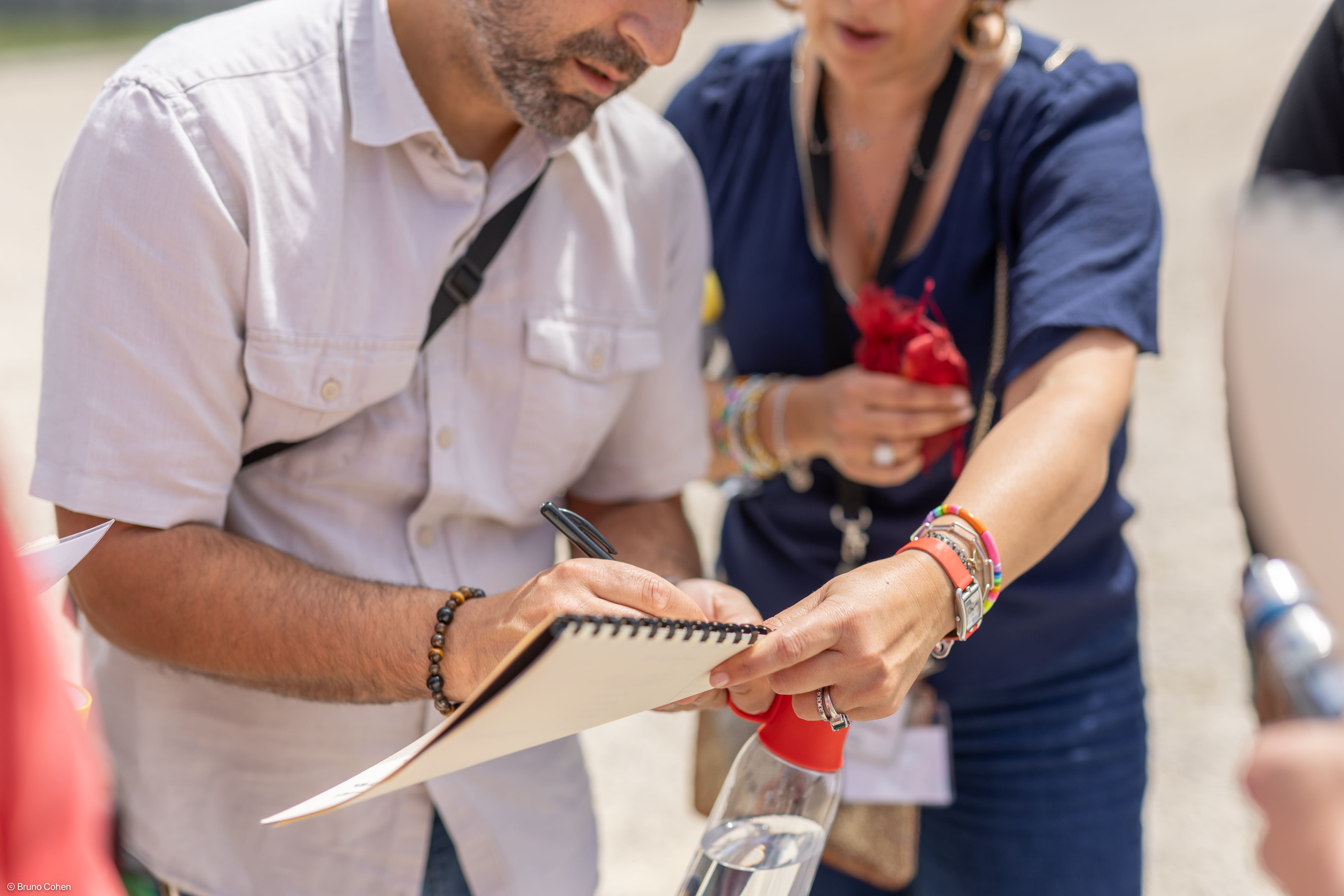 Man in white shirt writing on a spiral notebook held by a woman wearing a blue shirt and colorful bracelets.