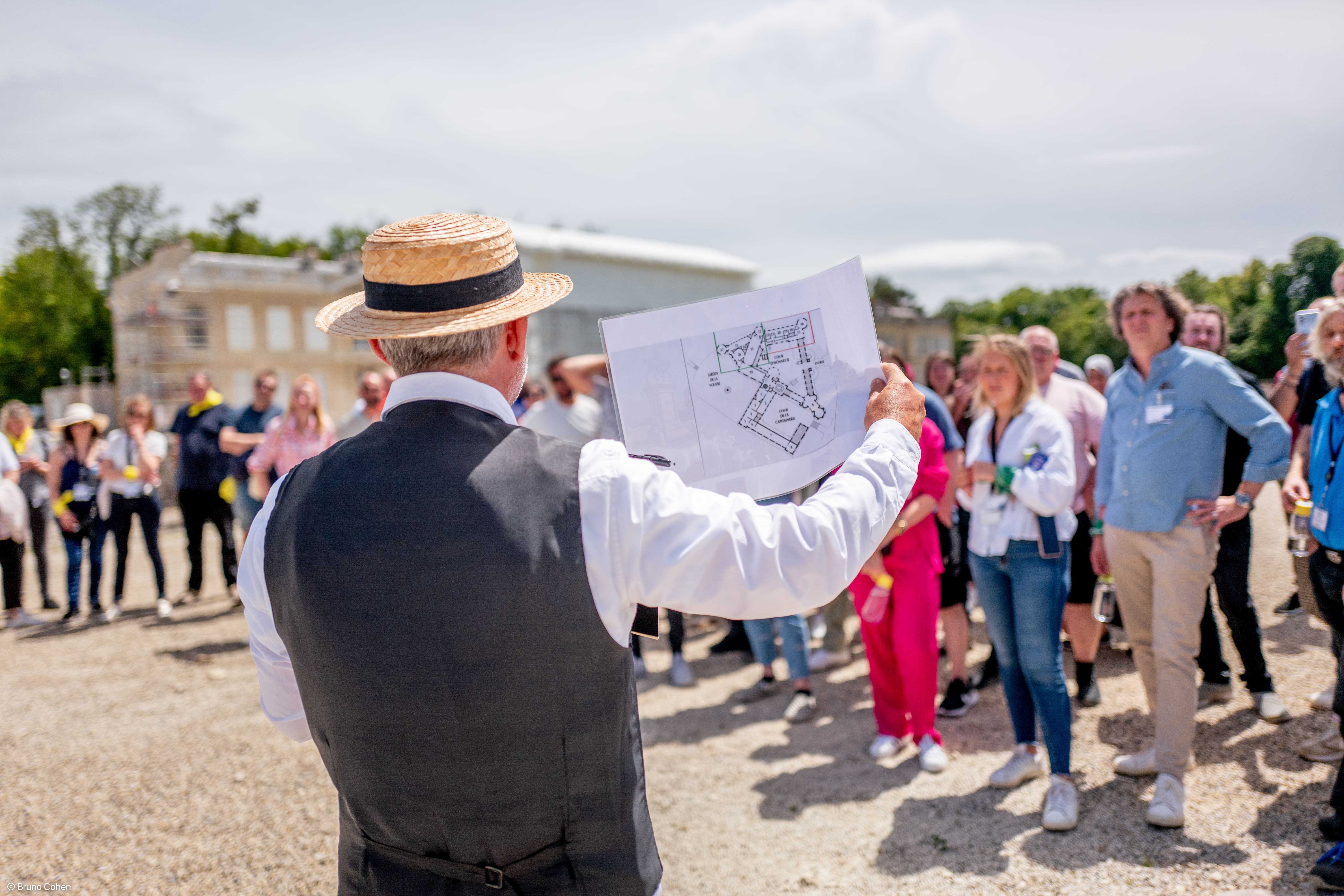 Man in a straw hat and vest holding a map while addressing a crowd outdoors near historic buildings.