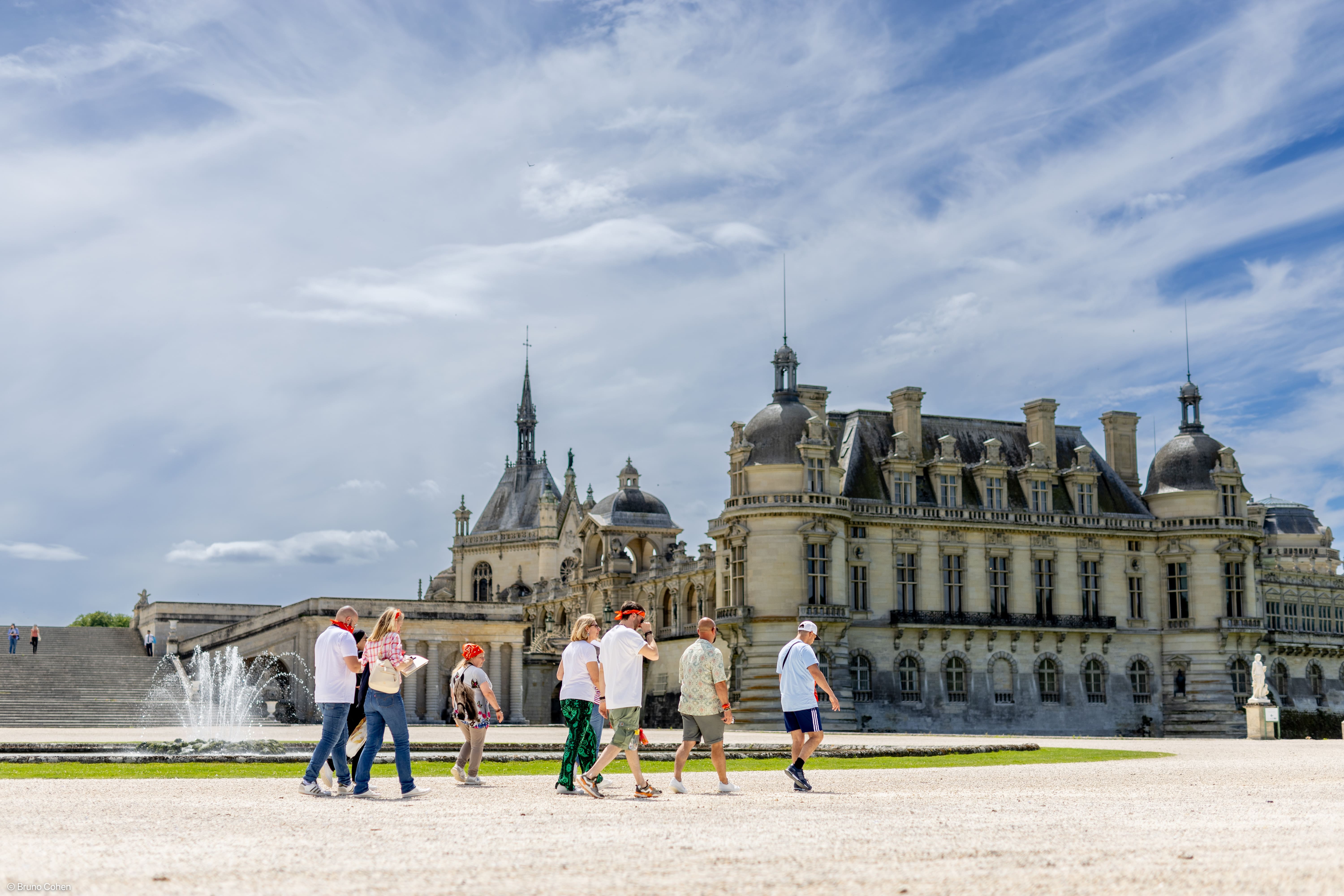 Group of tourists walking near a historic European castle with decorative architecture under a partly cloudy blue sky.