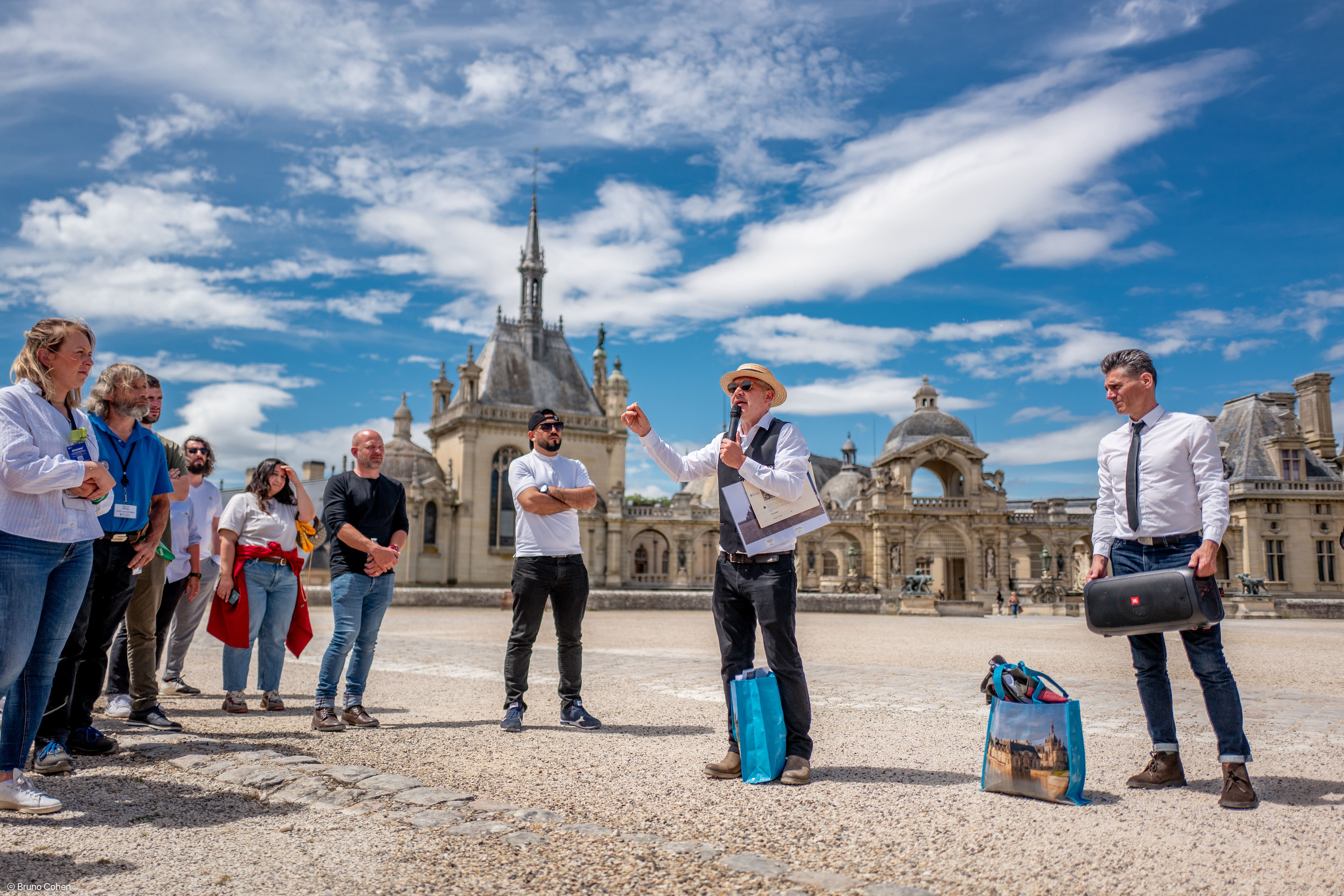 A group of people listening to a man speaking with a microphone outside a historic European castle under a blue sky with scattered clouds.