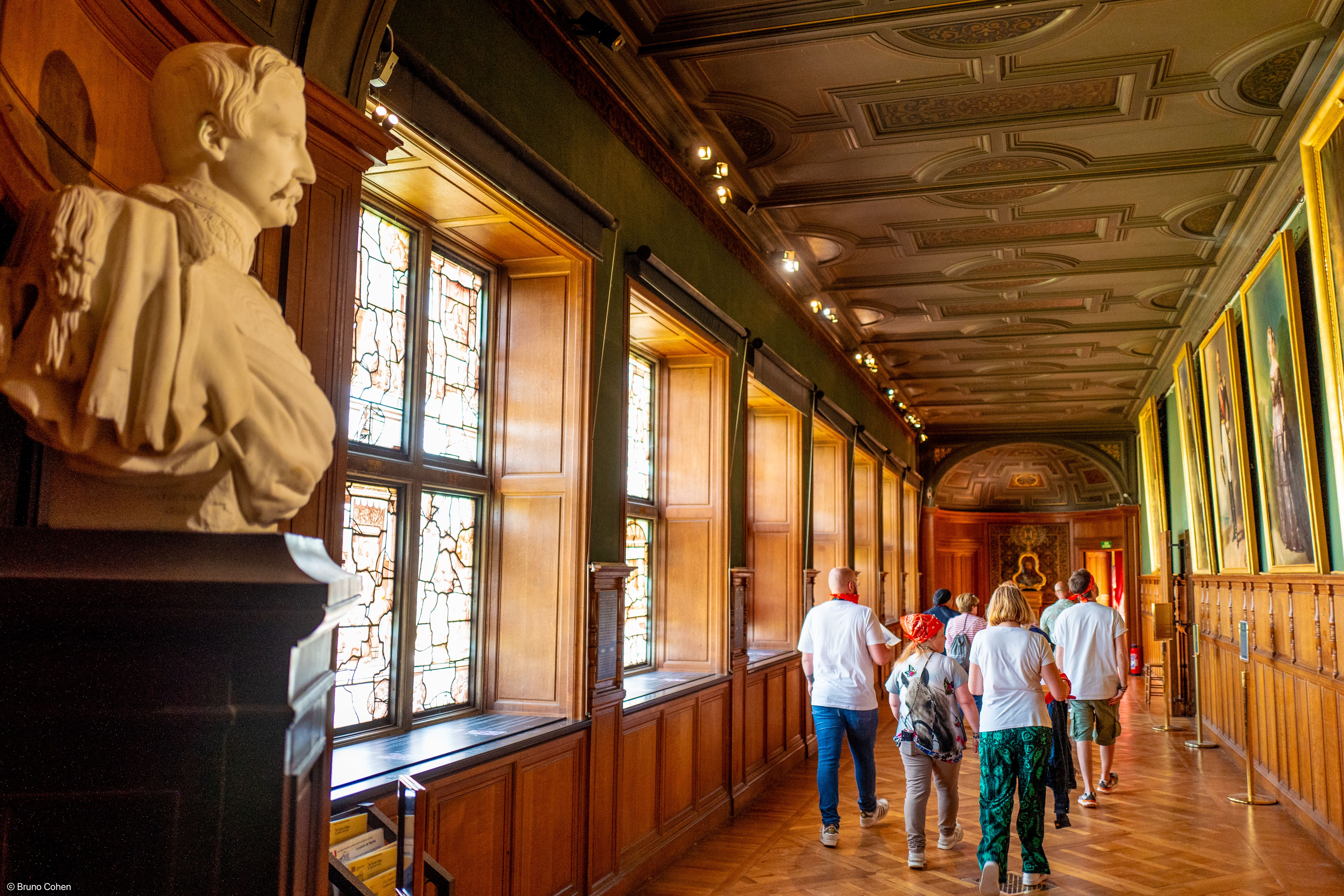 Group of people walking through a historic hall with wooden paneling, stained glass windows, a bust statue, and large framed portraits on the walls.