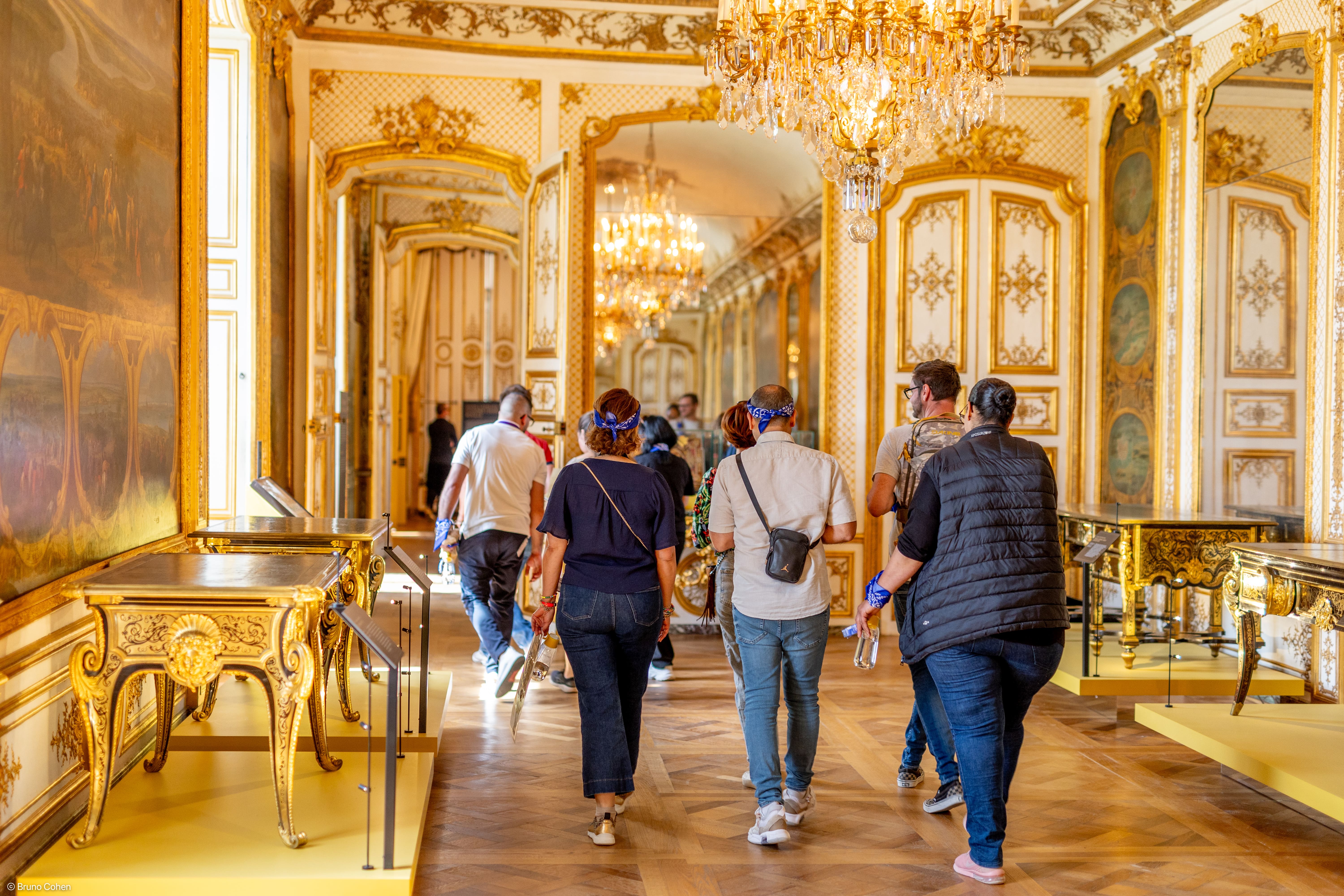 Group of tourists walking through a lavish museum hallway with golden ornate decorations and chandeliers.