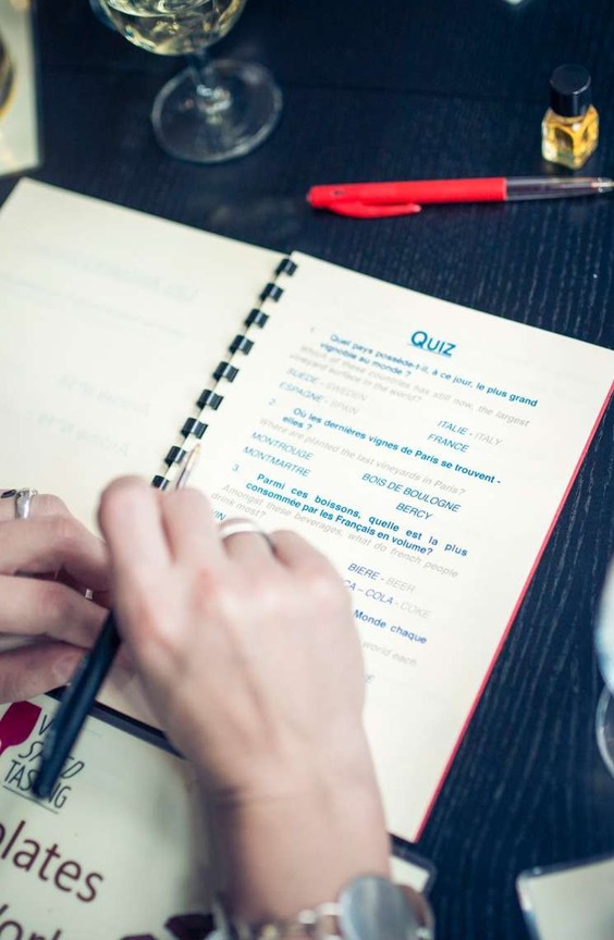 Person holding a pen and writing answers in a quiz booklet on a dark table with a glass and a red pen nearby.