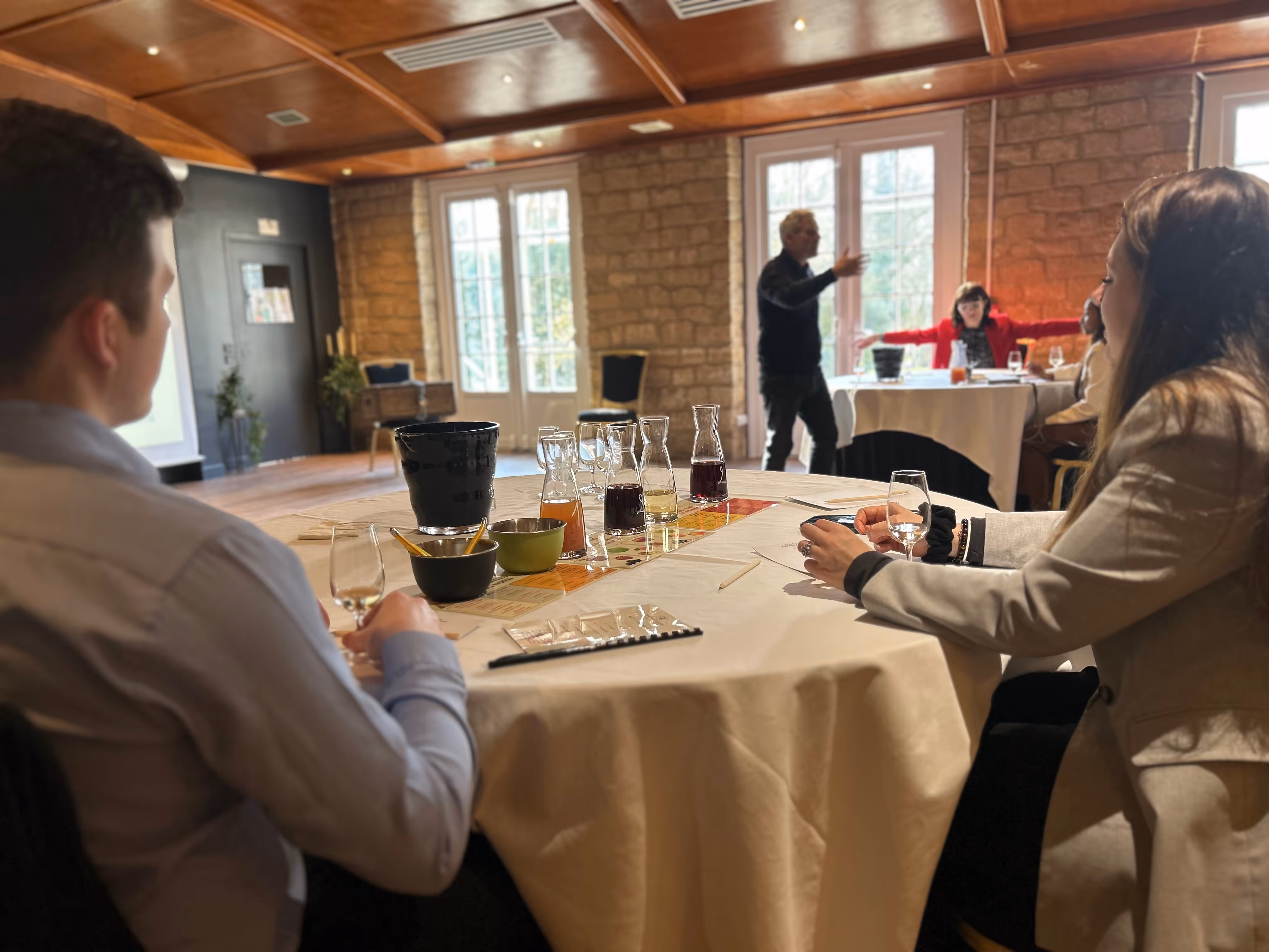 Two people seated at a table with wine glasses and carafes, listening to a person speaking in a sunlit room with stone walls.