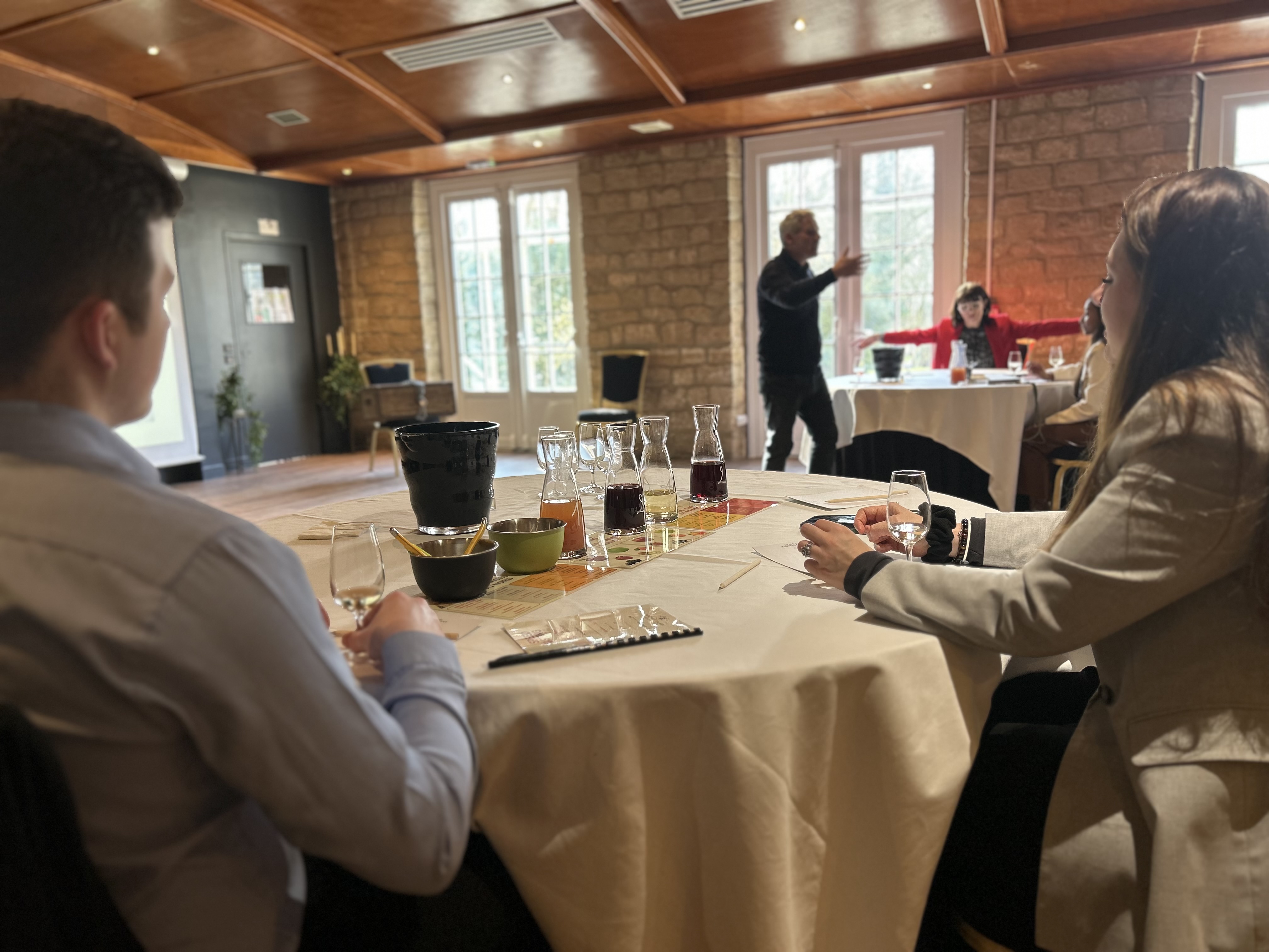 Two people seated at a table with wine glasses and carafes, listening to a person speaking in a sunlit room with stone walls.