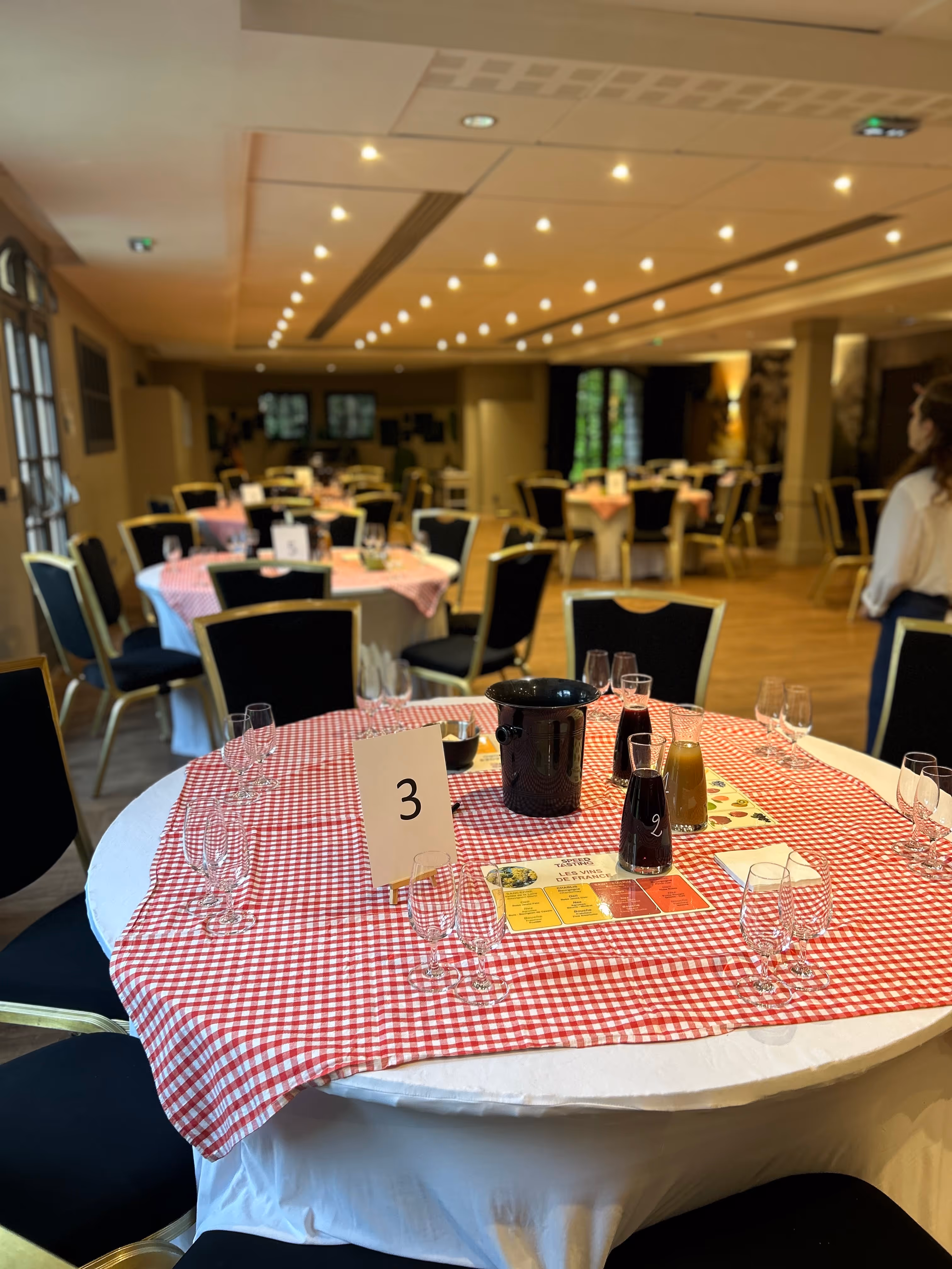 Round tables covered with red and white checkered cloths set up for wine tasting event with empty glasses, carafes with drinks, and a table number 3 card.