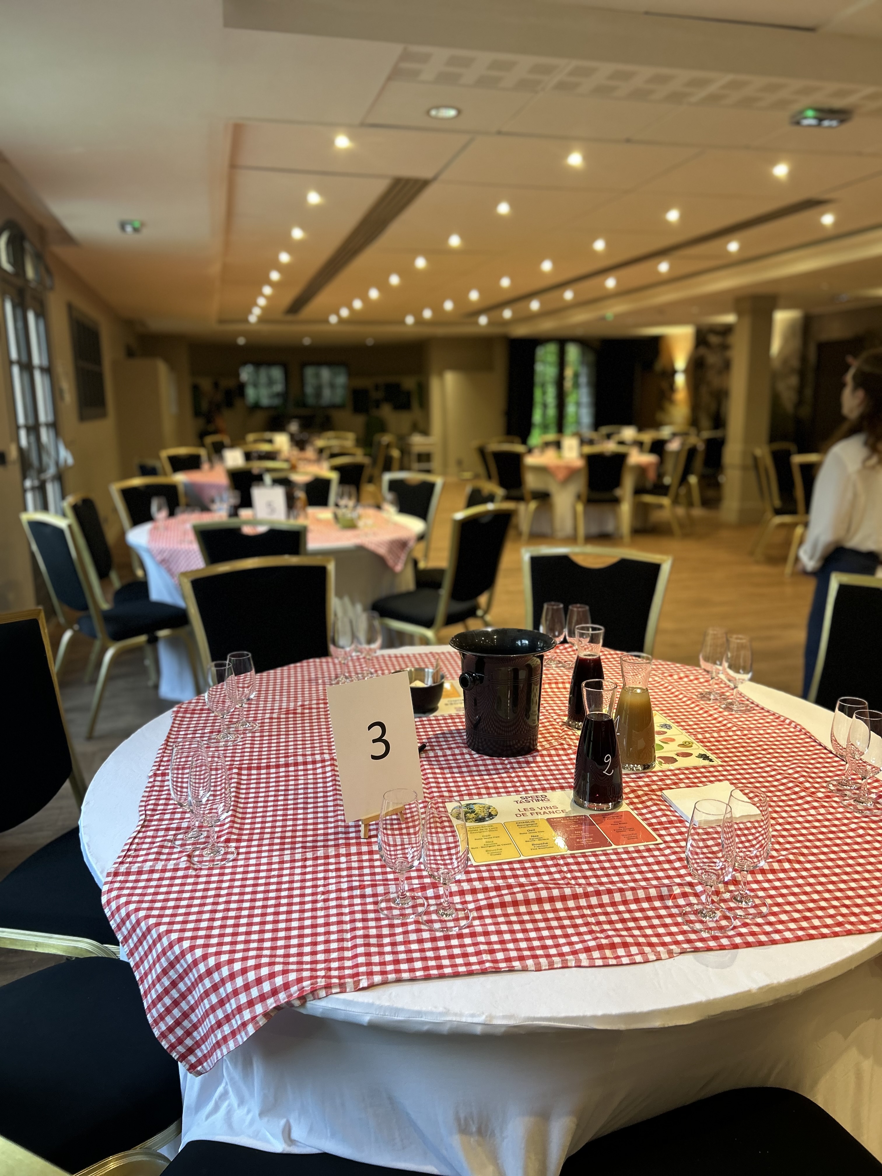 Round tables covered with red and white checkered cloths set up for wine tasting event with empty glasses, carafes with drinks, and a table number 3 card.