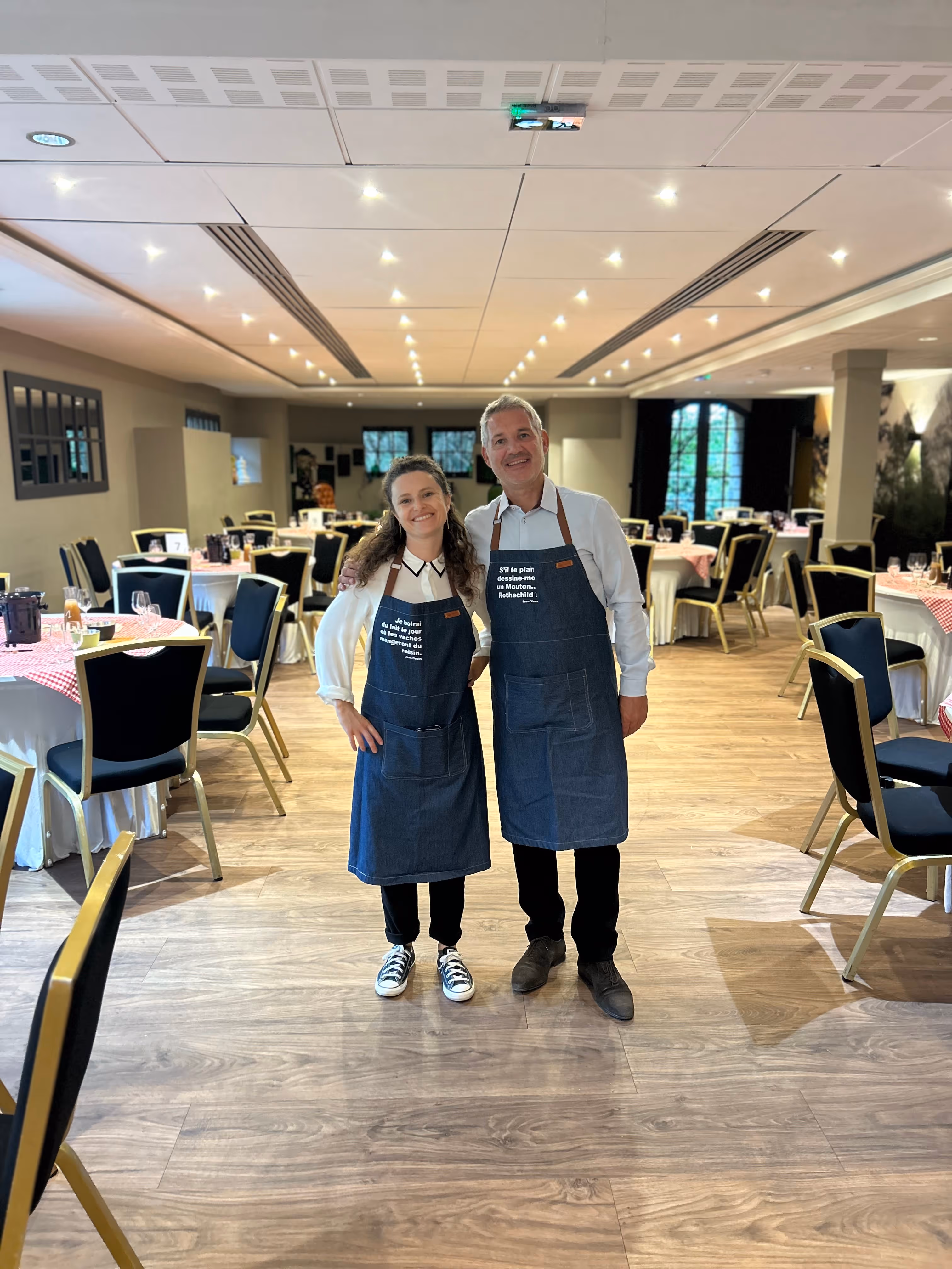 A smiling man and woman wearing denim aprons stand arm in arm in a warmly lit, empty restaurant dining room with round tables covered in white and red checkered tablecloths.