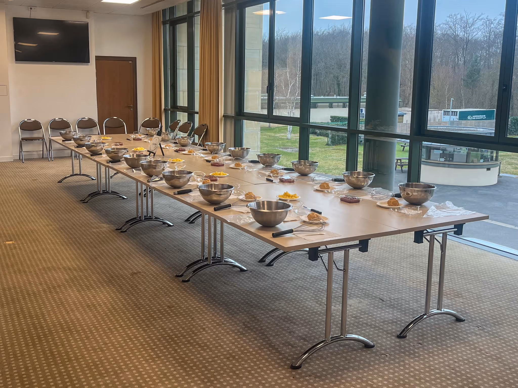 Long table set for a cooking class with metal mixing bowls, whisks, plates of ingredients, and chairs in a bright room with large windows.