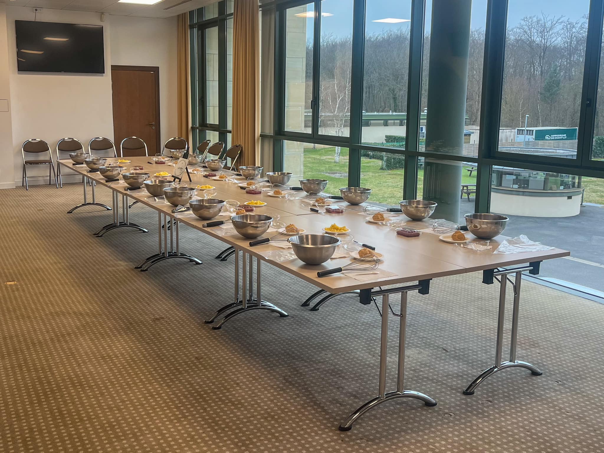 Long table set for a cooking class with metal mixing bowls, whisks, plates of ingredients, and chairs in a bright room with large windows.