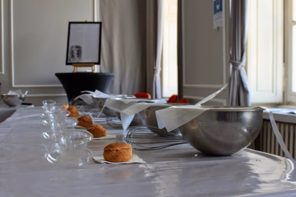 Row of baking bowls, whisks, glass bowls, and cream puffs arranged on a table in a bright room with large windows.