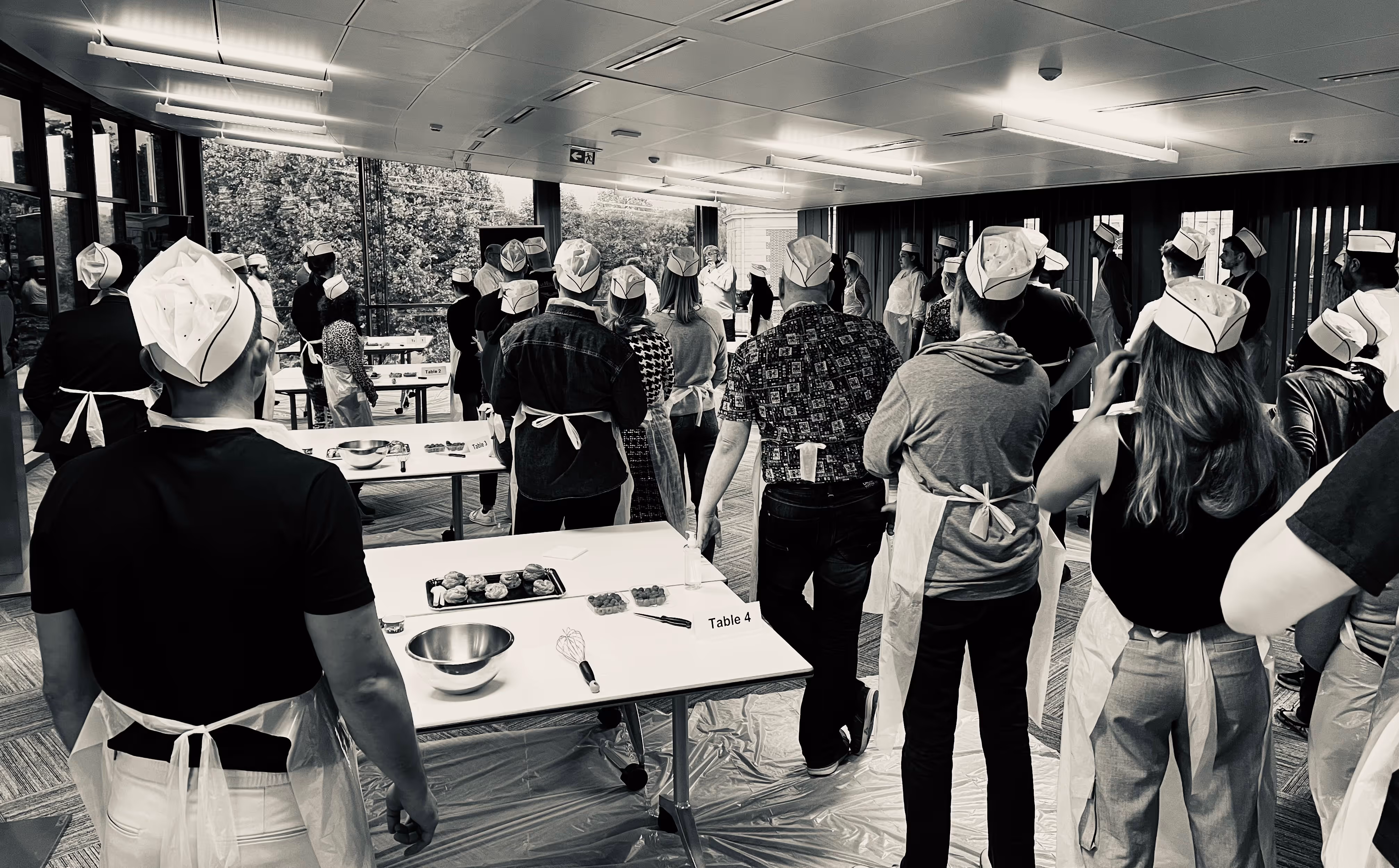 Group of people wearing chef hats and aprons standing around tables set with mixing bowls, whisks, and pastries in a bright room.