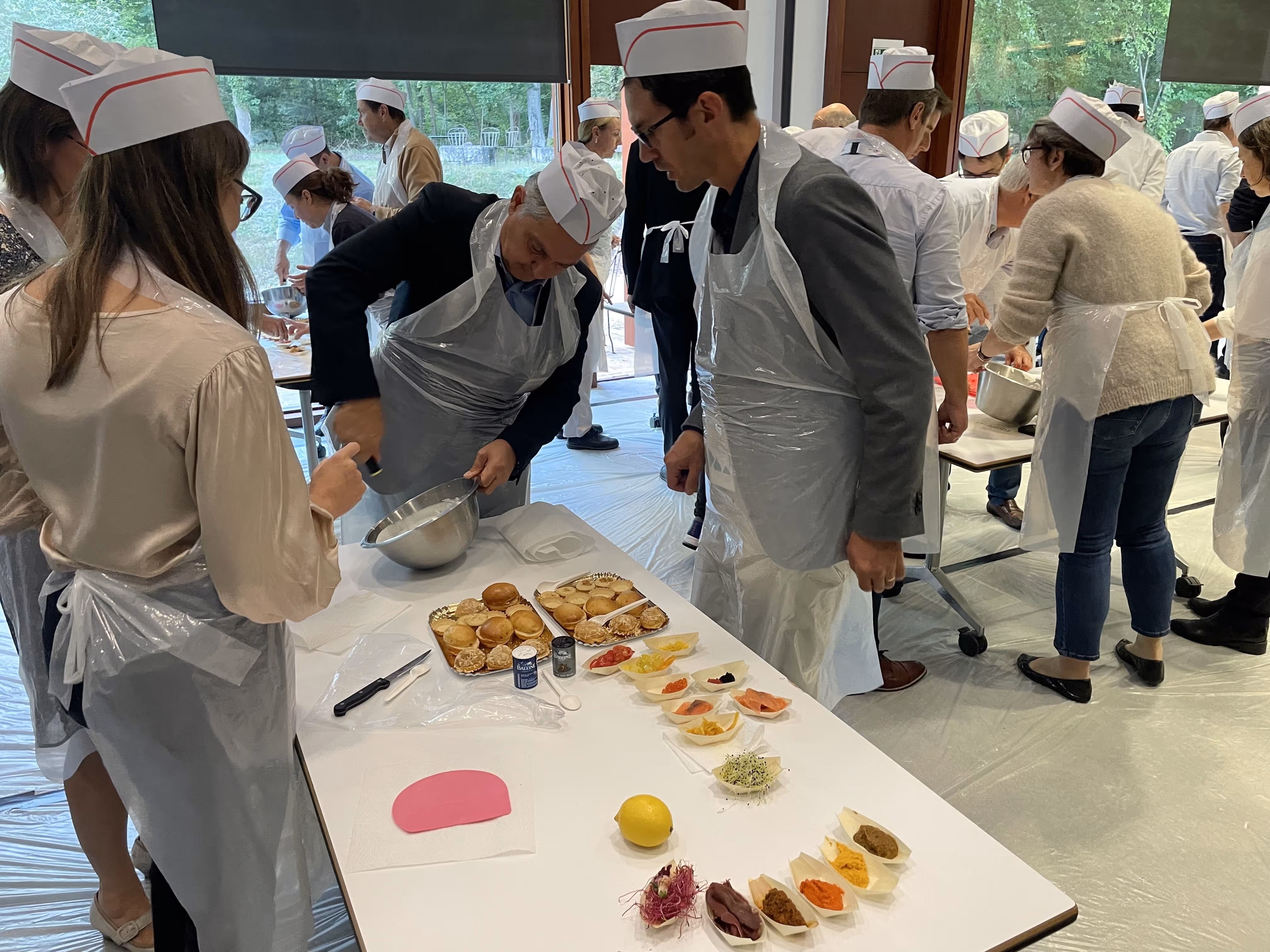 Group of people wearing white hats and aprons taking a cooking class, mixing ingredients and preparing food on tables.