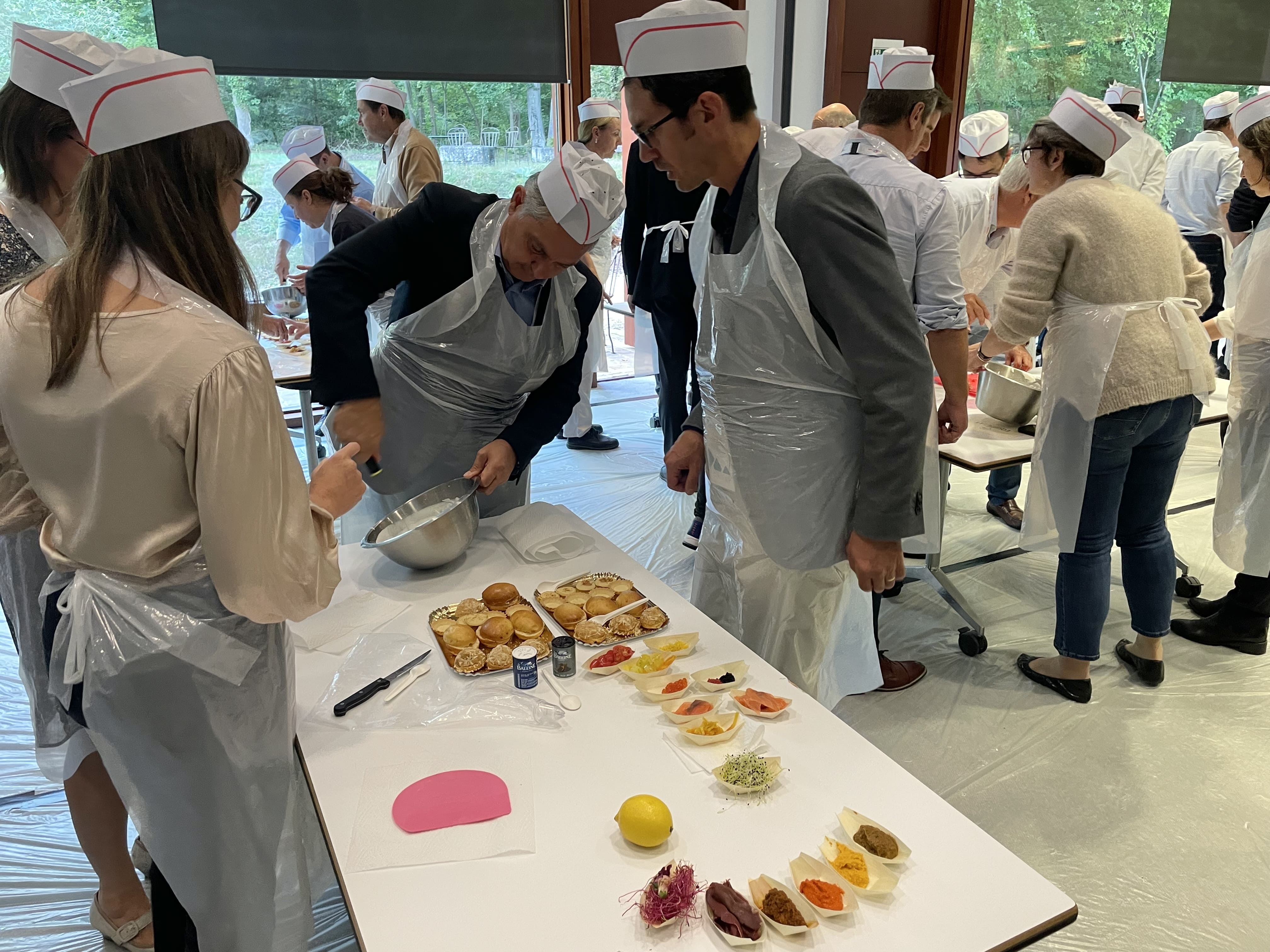 Group of people wearing white hats and aprons taking a cooking class, mixing ingredients and preparing food on tables.