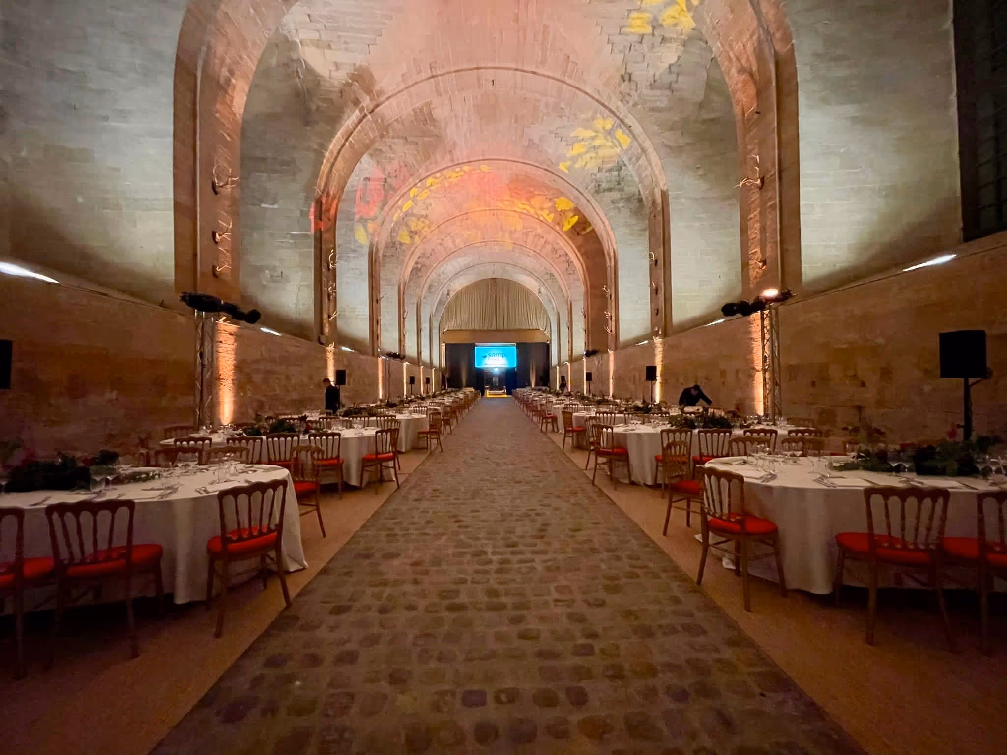 Long stone hall with arched ceiling set up for a banquet with round tables and red chairs arranged on both sides.