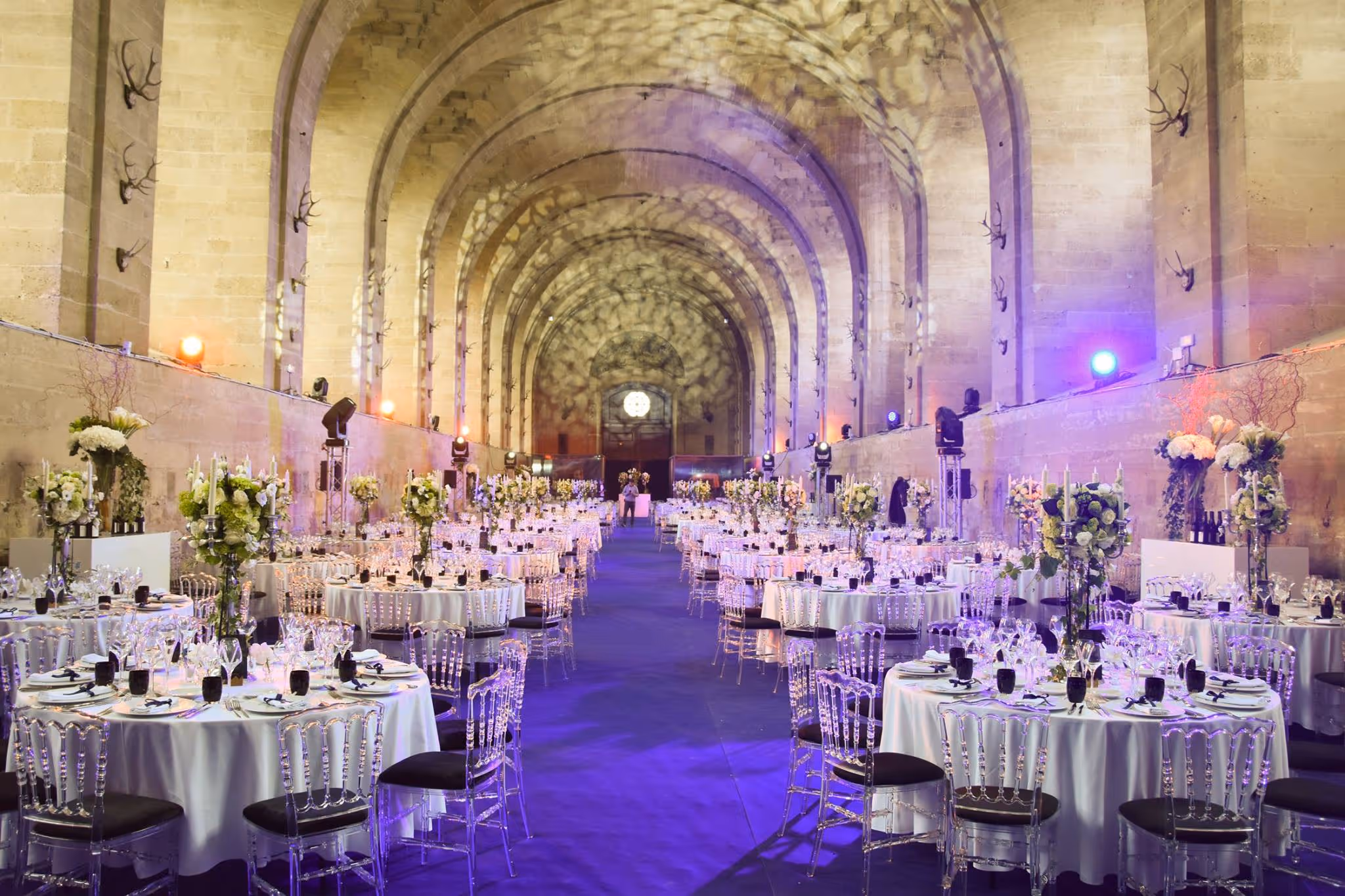 Large banquet hall with high arched ceilings, decorated tables with white cloths, clear chairs, and floral centerpieces under soft purple lighting.