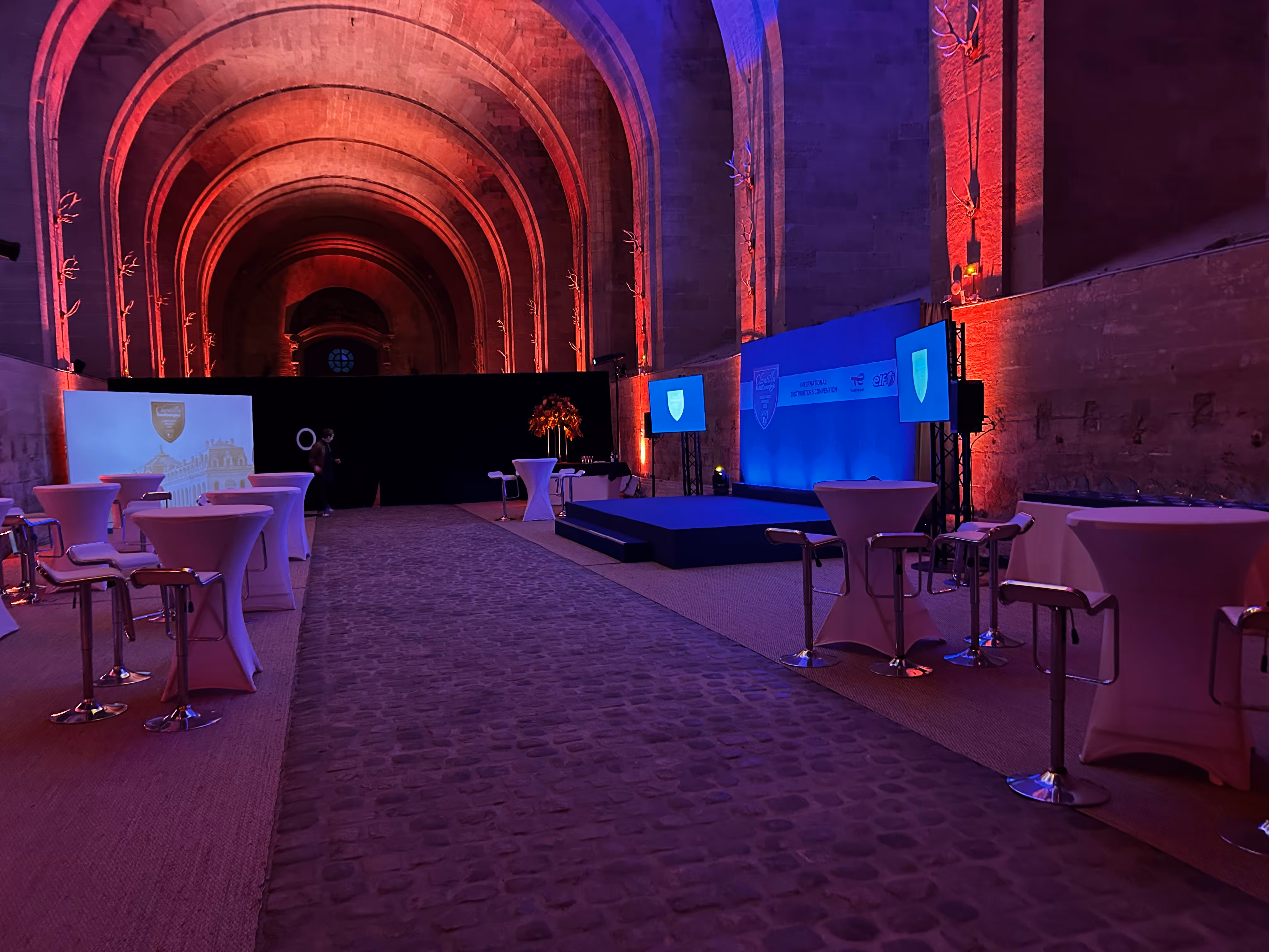 Event setup inside a large historic stone hall with arched ceilings, featuring a stage with blue screens, red uplighting, and tall cocktail tables with white covers and bar stools.