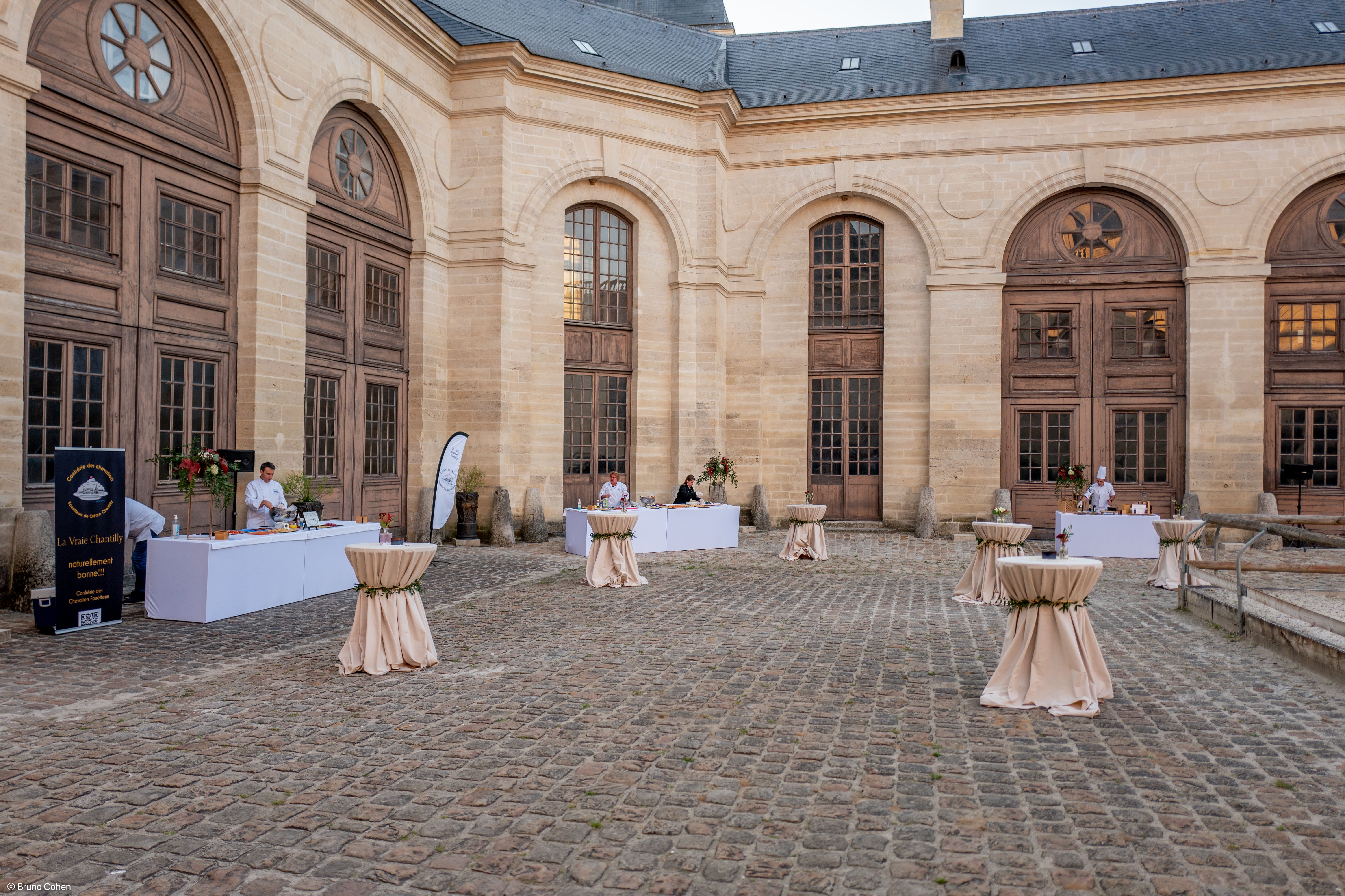 Outdoor event setup with chefs standing behind white tables and round cocktail tables draped with beige cloth in front of large arched wooden doors.