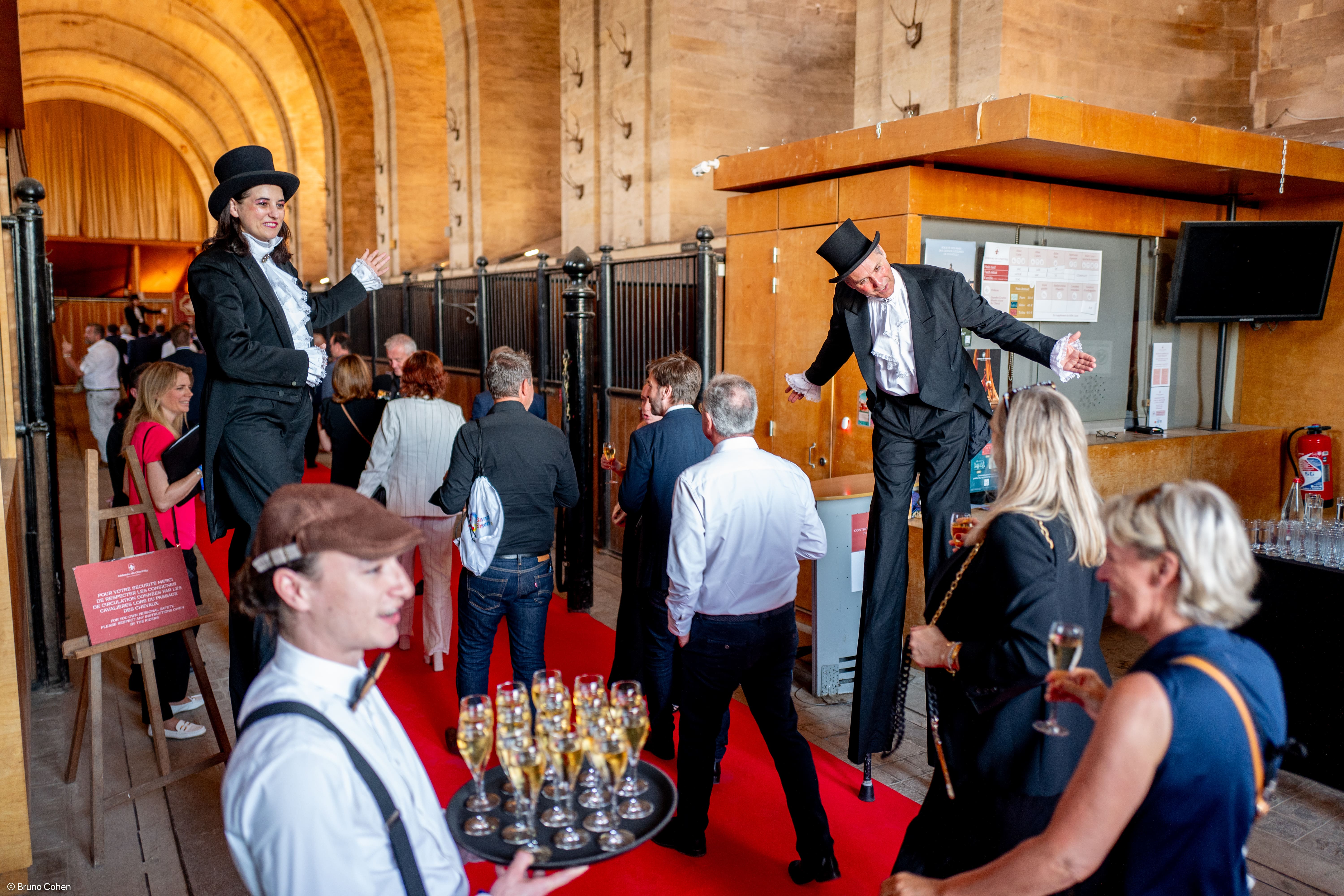 People walking on a red carpet inside a building with two performers in top hats and tails greeting guests and a waiter holding a tray of champagne glasses.