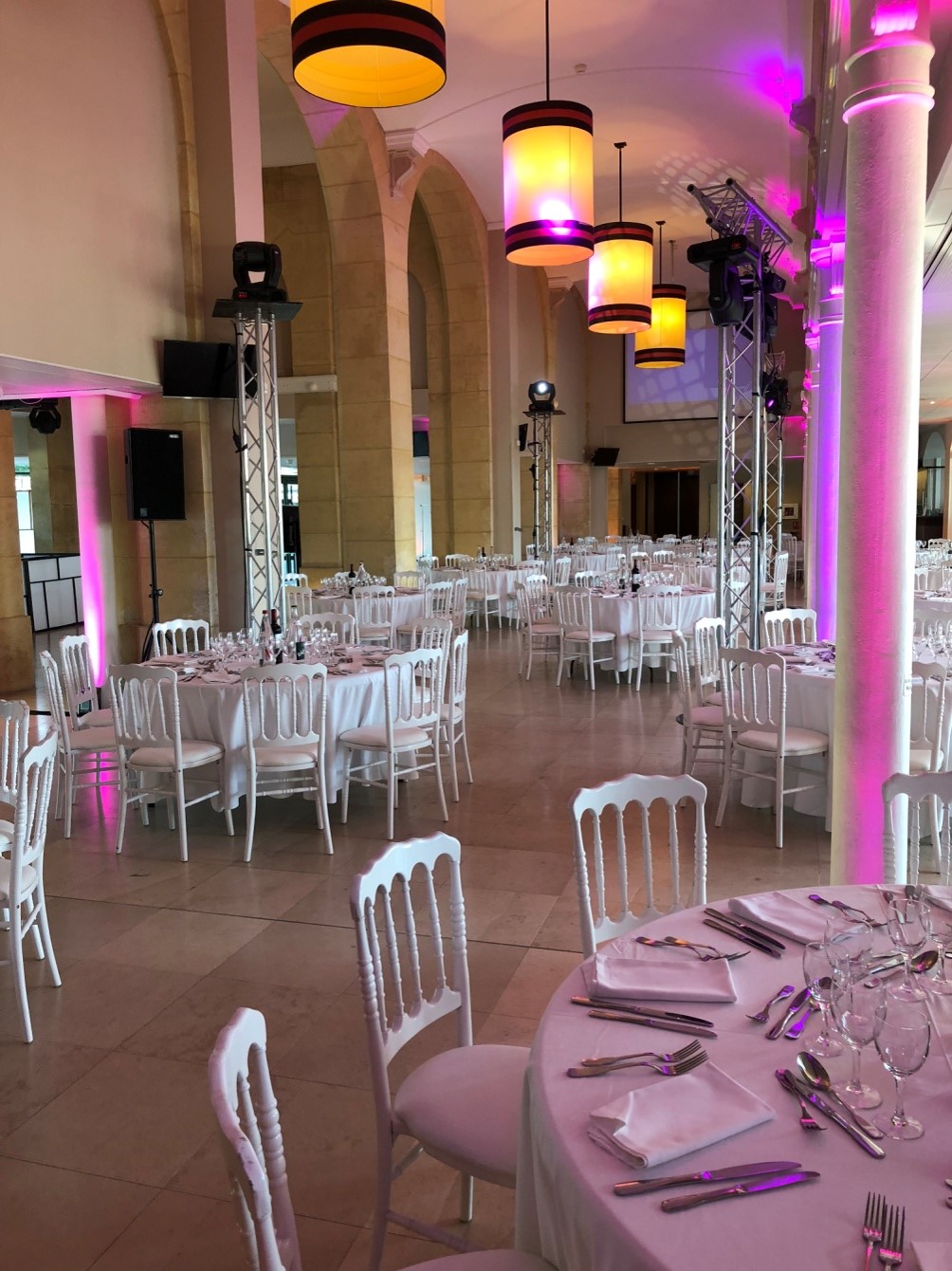 Elegant banquet hall with round tables draped in white linens, set with silverware, glassware, and napkins, lit by warm hanging lamps and purple uplighting on columns.