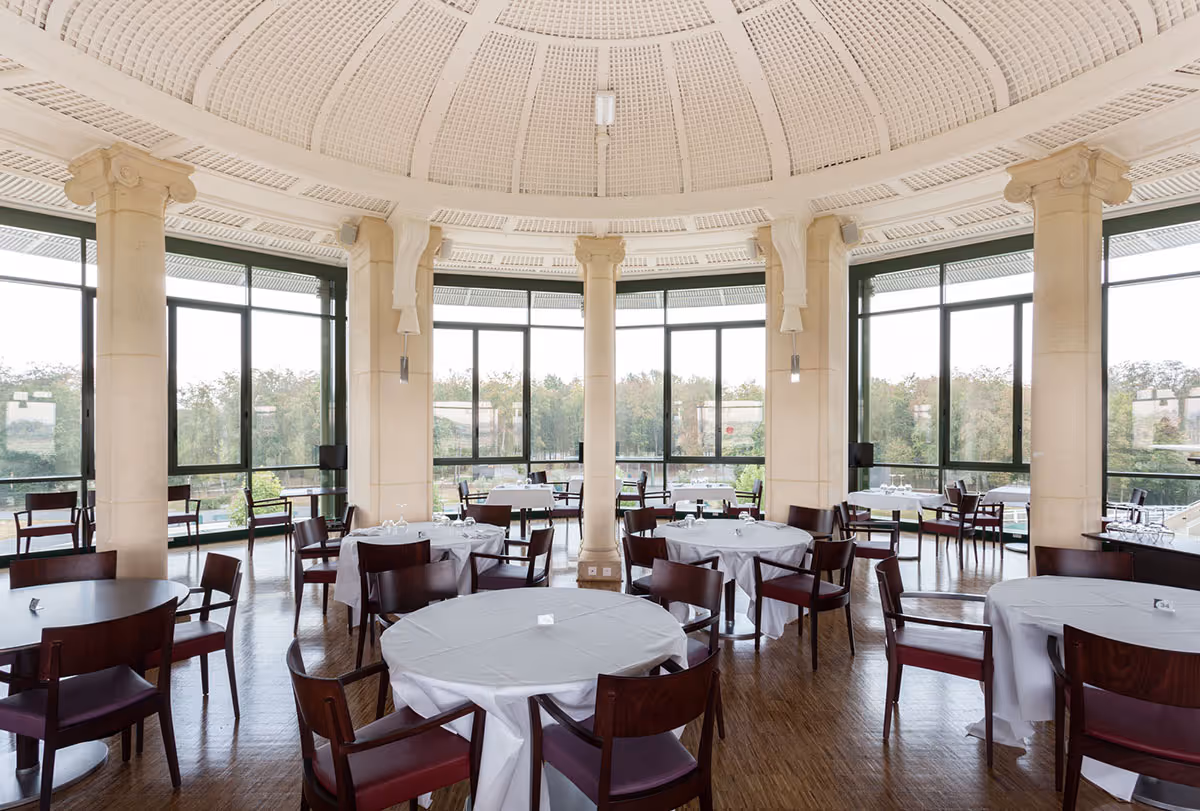 Spacious dining room with round tables covered in white tablecloths and dark wood chairs under a large domed ceiling with tall windows showing trees outside.