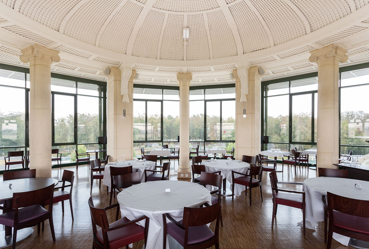 Spacious dining room with round tables covered in white tablecloths and dark wood chairs under a large domed ceiling with tall windows showing trees outside.