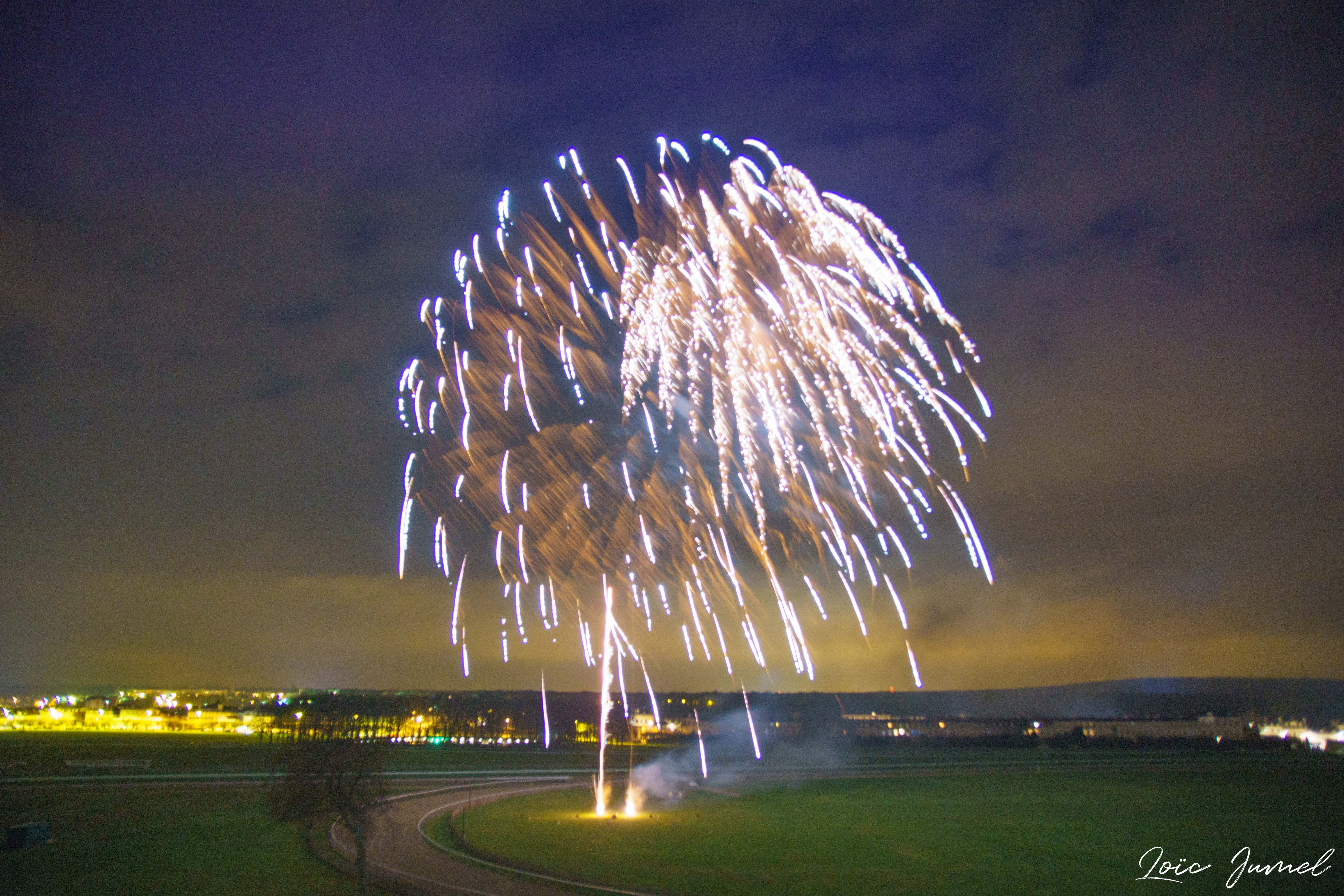 Bright fireworks bursting over a curved road and green field at night with city lights in the background.