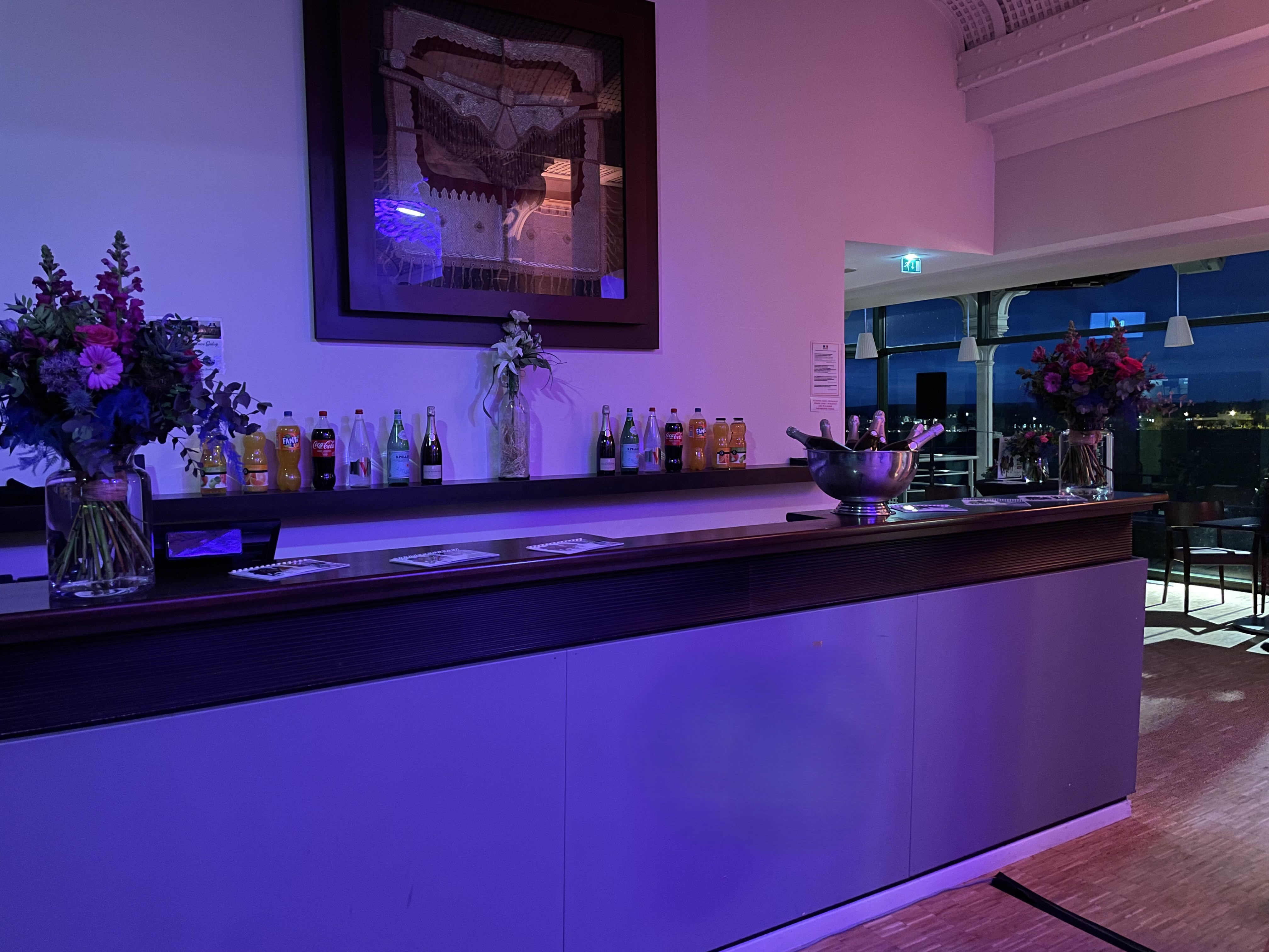 Bar counter with assorted bottled drinks, two large floral arrangements, and a metal ice bucket with champagne bottles under purple lighting in an indoor event space.