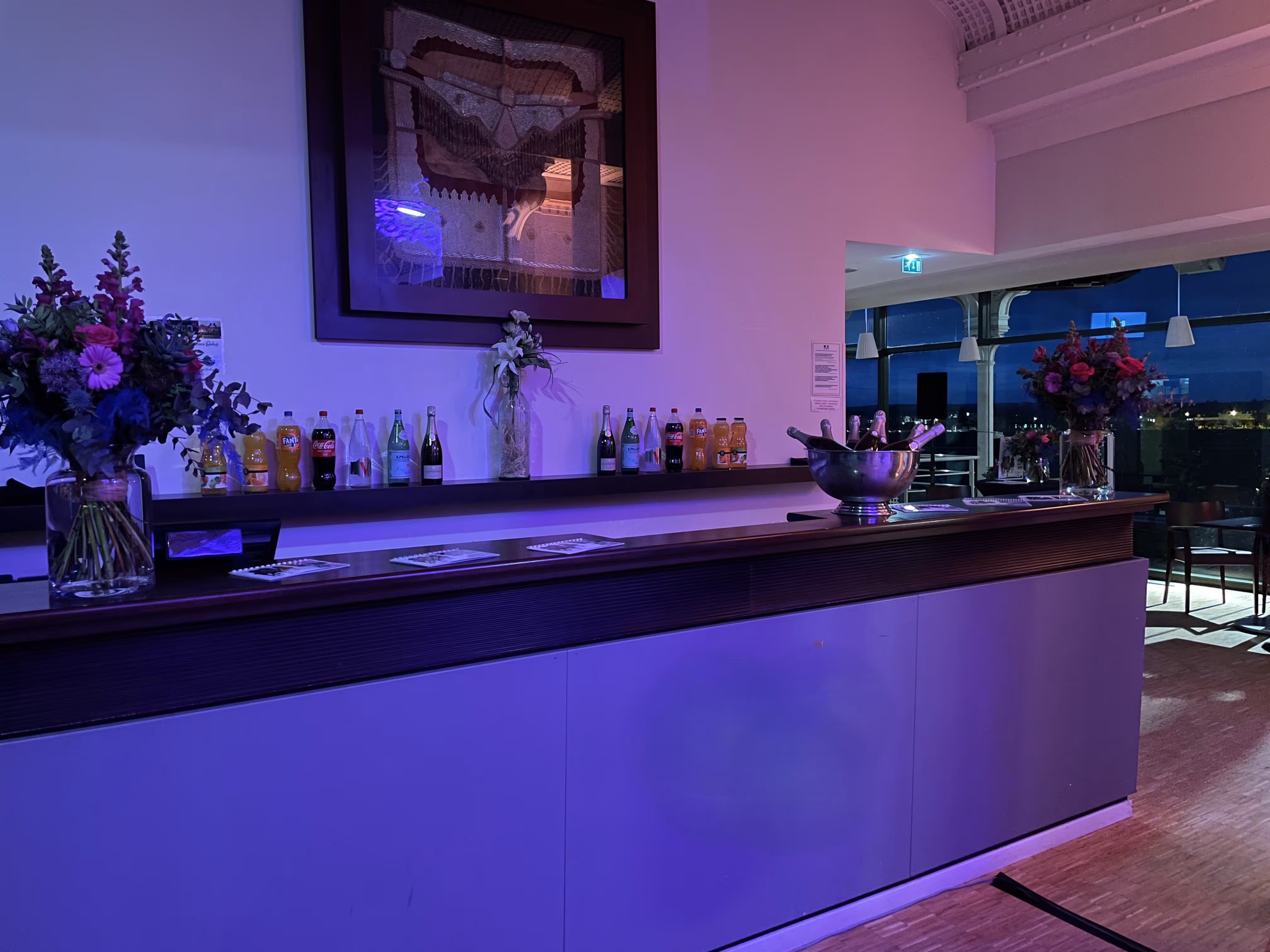 Bar counter with assorted bottled drinks, two large floral arrangements, and a metal ice bucket with champagne bottles under purple lighting in an indoor event space.