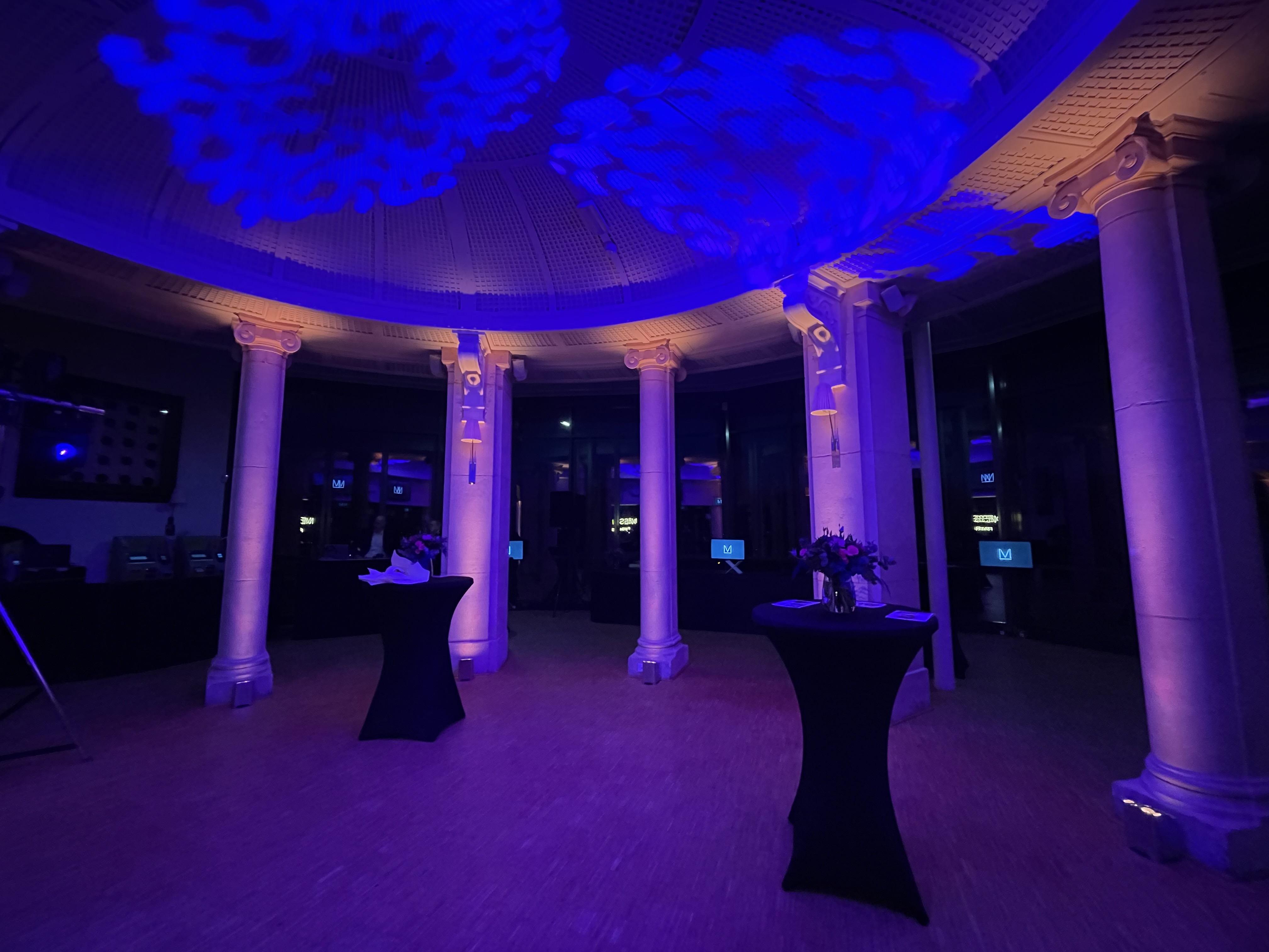 Elegant circular room with illuminated classical columns, blue decorative ceiling lights, and two tall black cocktail tables with floral arrangements.