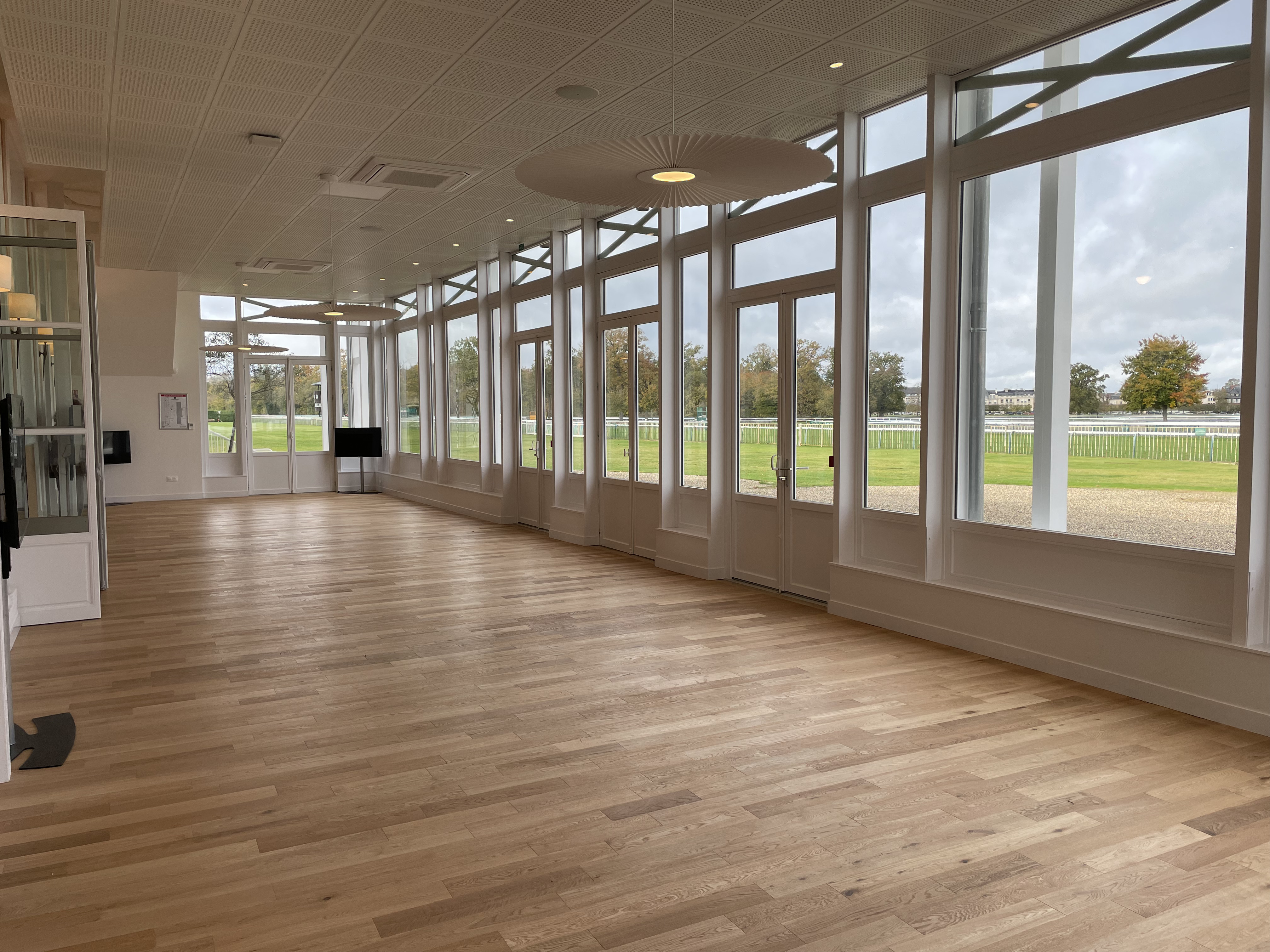 Empty spacious room with wooden floor and large windows overlooking a grassy outdoor area and trees under a cloudy sky.